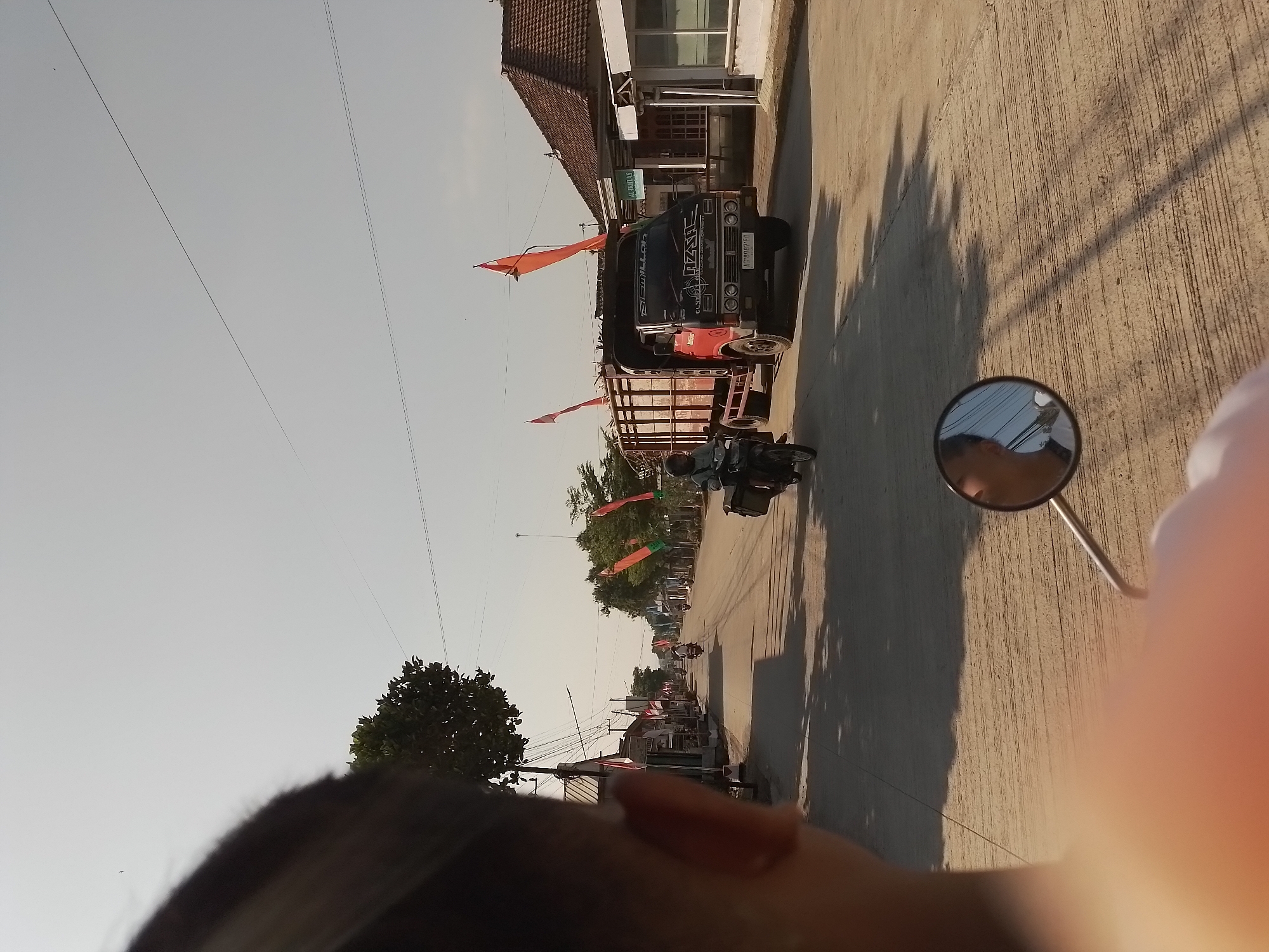 The image depicts a street scene in Ngancar, Indonesia, captured from the perspective of someone riding a motorcycle. The foreground is dominated by the head and shoulder of the rider, partially obscuring the view ahead. In the middle ground, a red truck with a wooden cargo bed is driving down the street, adorned with two orange flags. A motorcycle is traveling ahead of the truck, with a rider visible. Various buildings line the left side of the street, appearing to be shops or residences, many with awnings. Orange flags are also displayed along the sides of the road, suggesting a local celebration or event. The street itself is paved with concrete and shows visible lines. The right side of the street features more buildings, including one with a tiled roof and a sign that reads "AL IKHLAS". The overall environment suggests a warm, sunny day in a rural or semi-urban Indonesian setting.