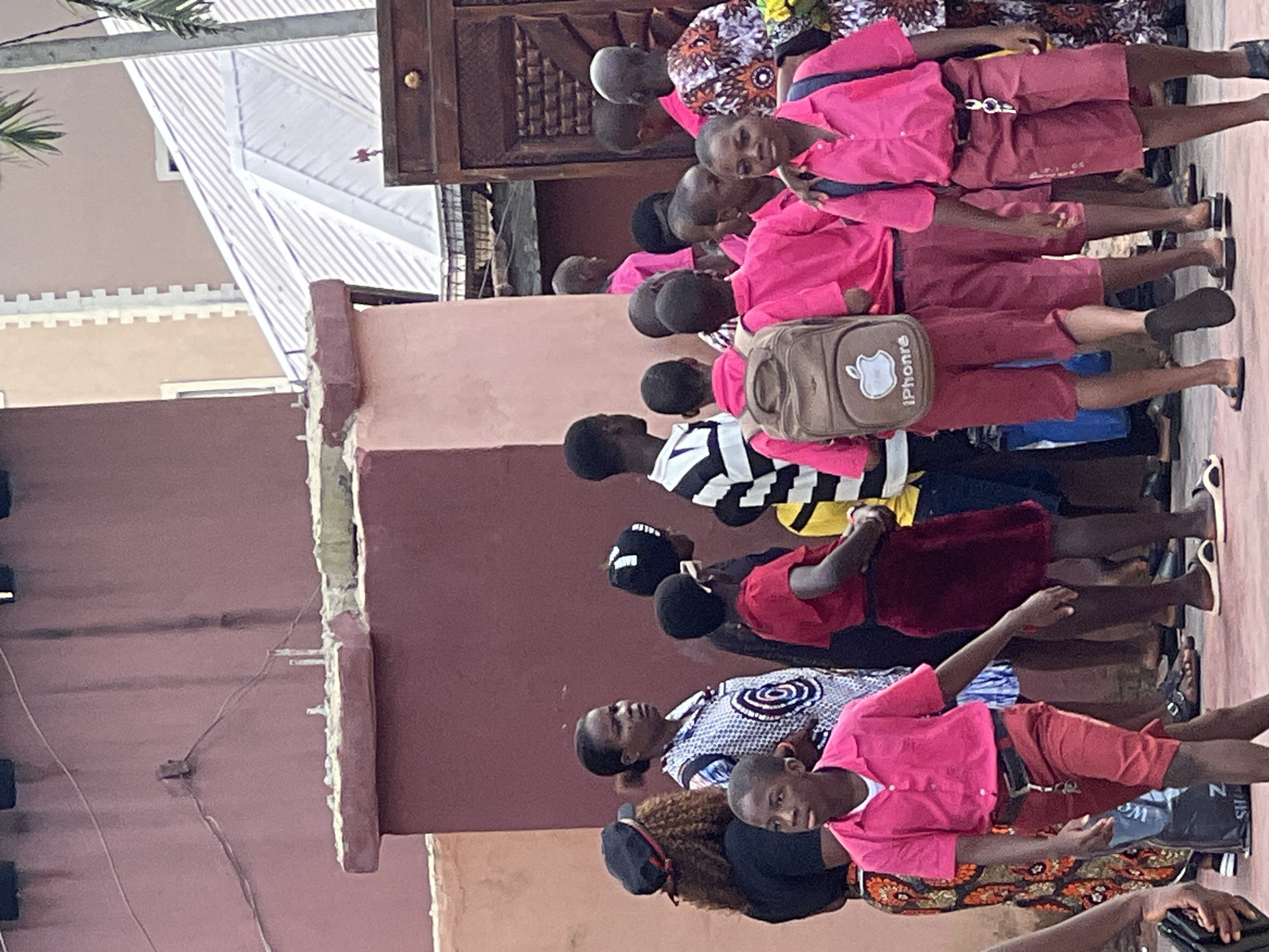 This image captures a group of people, primarily children in school uniforms, gathered on a street in Nnewi, Nigeria. The scene appears to be outside a building with a textured, reddish-brown wall. The children are mostly wearing pink shirts and red shorts or skirts, suggesting they are students. Some are carrying backpacks, one of which prominently displays an Apple logo with the text "iPhone". The environment includes a tall, thin tree with palm-like fronds on the right side, and a corrugated metal roof of a structure in the background. The lighting suggests it is daytime. The overall impression is a common street scene in a Nigerian city, possibly during school dismissal or an event.