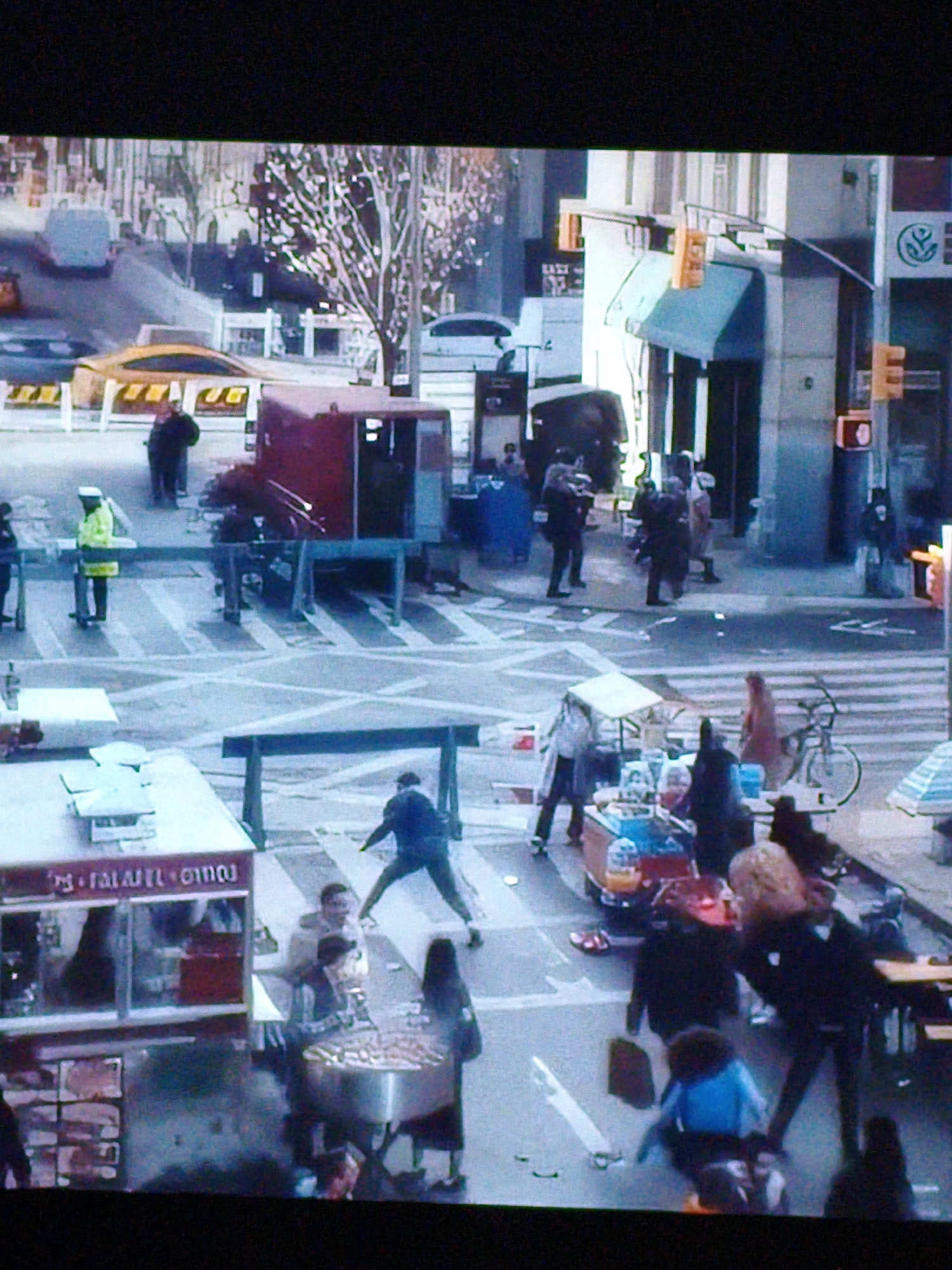 This is a ground-level, medium-distance, street-level shot of a busy urban environment that appears to be a food cart district or marketplace. The scene is packed with people and food vendors. In the foreground, a food cart labeled with what appears to be "Falafel" in Arabic script is prominent. A vendor is serving customers from this cart. To the left, another food vendor setup is visible. A few feet away, a group of people are gathered, some interacting with the vendors, while others are standing or walking. In the background, several other food carts and stalls are visible, along with more people milling about. There are a few vehicles in the background, including what appears to be a delivery truck. Traffic lights and street signs are visible, suggesting a public street. The overall atmosphere is one of a bustling market or street fair. There is no indication this scene is located in Vladivostok, Russia, and there are no distinctive architectural or environmental cues to support that claim.