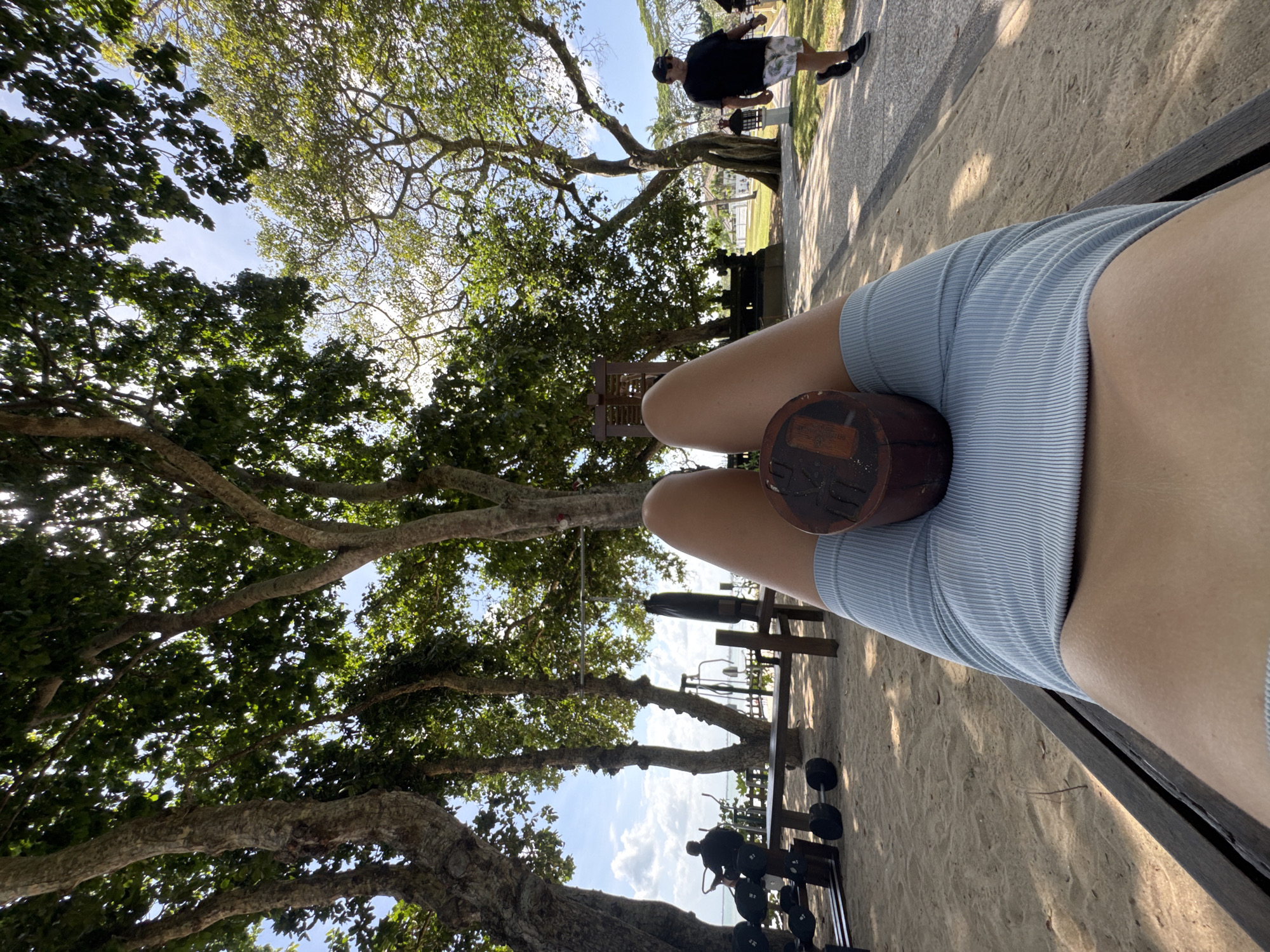 An overhead, first-person perspective shot shows a person's legs, clad in light blue ribbed shorts, resting on a wooden bench. A cylindrical wooden object rests on the person's thighs. The bench is situated on a sandy area beneath the dappled shade of large, mature trees. In the background, outdoor exercise equipment, including weight racks with dumbbells and a punching bag, are visible. Further back, a person in a black t-shirt and patterned shorts walks along a paved pathway. The scene is set in an outdoor gym or park area, likely in Kuta Selatan, Indonesia, given the presence of tropical trees and the overall ambiance. The sky is bright blue with scattered white clouds.