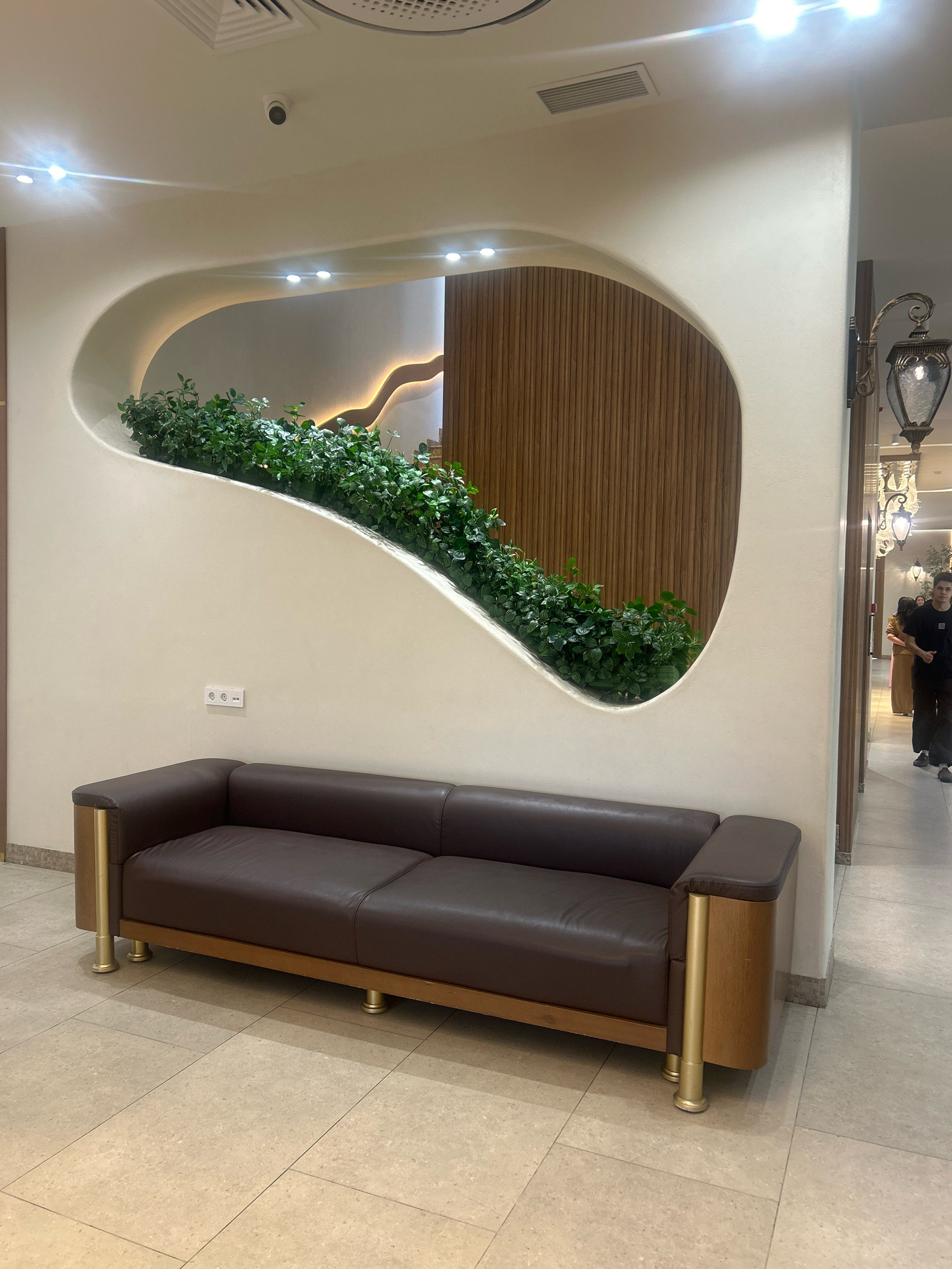 A dark brown leather sofa is positioned in front of a wall with a large, organically shaped recess filled with lush green plants. The recess is illuminated by soft lighting, highlighting the foliage and a wooden slatted panel behind it. The wall itself is a light cream color, with a subtle textured finish. Two electrical outlets are visible on the wall above the sofa. The floor is tiled with large, light-colored square tiles. To the right of the sofa and wall feature, a hallway is visible, with decorative lamps and several people walking. One person, a young man in a black t-shirt and dark pants, is walking towards the foreground. The scene appears to be an interior space, possibly a lobby or waiting area, in Almaty, Kazakhstan.