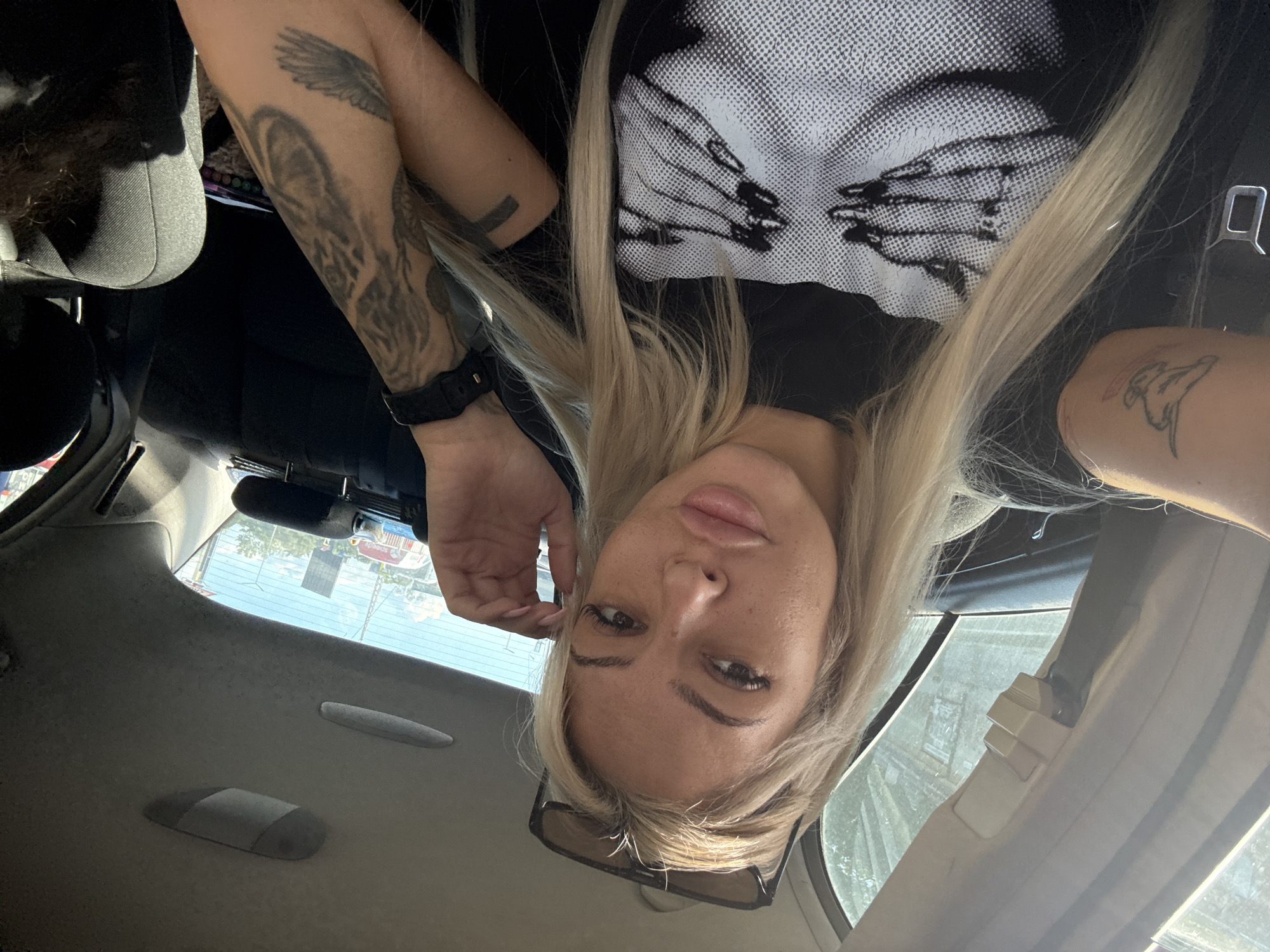 A medium, inverted, selfie-style shot depicts a fair-skinned woman with blonde hair inside a car. She is wearing a black t-shirt with a white graphic design of two hands on the front. Her arms are visible, with tattoos on both forearms. The tattoo on her left forearm appears to include a bird wing and a wolf-like animal, while the right forearm shows a smaller, minimalist design. She wears a black wristwatch.
The car's interior, including the ceiling, seats, and seatbelts, is visible. A pair of sunglasses rests partially below her face. Through the car window, glimpses of an urban setting are visible, but without sufficient detail to confirm the location as Plovdiv, Bulgaria.
