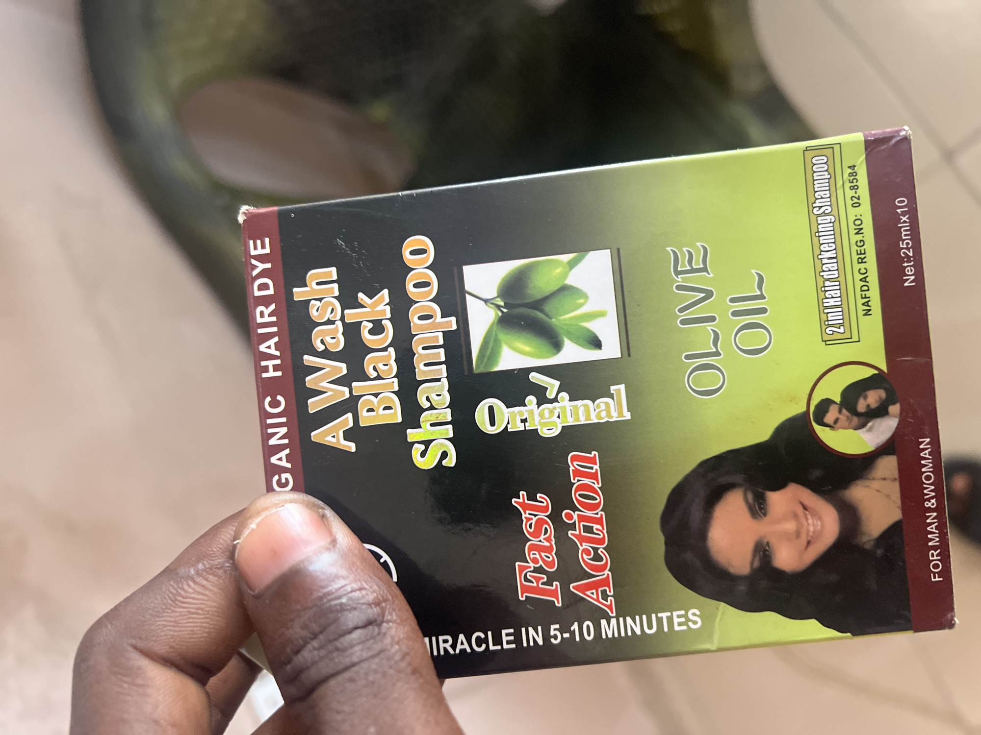 The image shows a hand holding a box of "A Wash Black Shampoo" for hair dye, with "Organic Hair Dye" written above. The box features an image of olives and a woman's face. The text "Fast Action" and "Original" are prominent. The box also indicates "Olive Oil" and "2in1 Hair darkening Shampoo" with a NAFDAC registration number. At the bottom, it states "For Man & Woman" and "Net: 25mlx10". The background is blurred, but appears to be an indoor setting with a green surface. The image was captured in Jalingo, Nigeria.