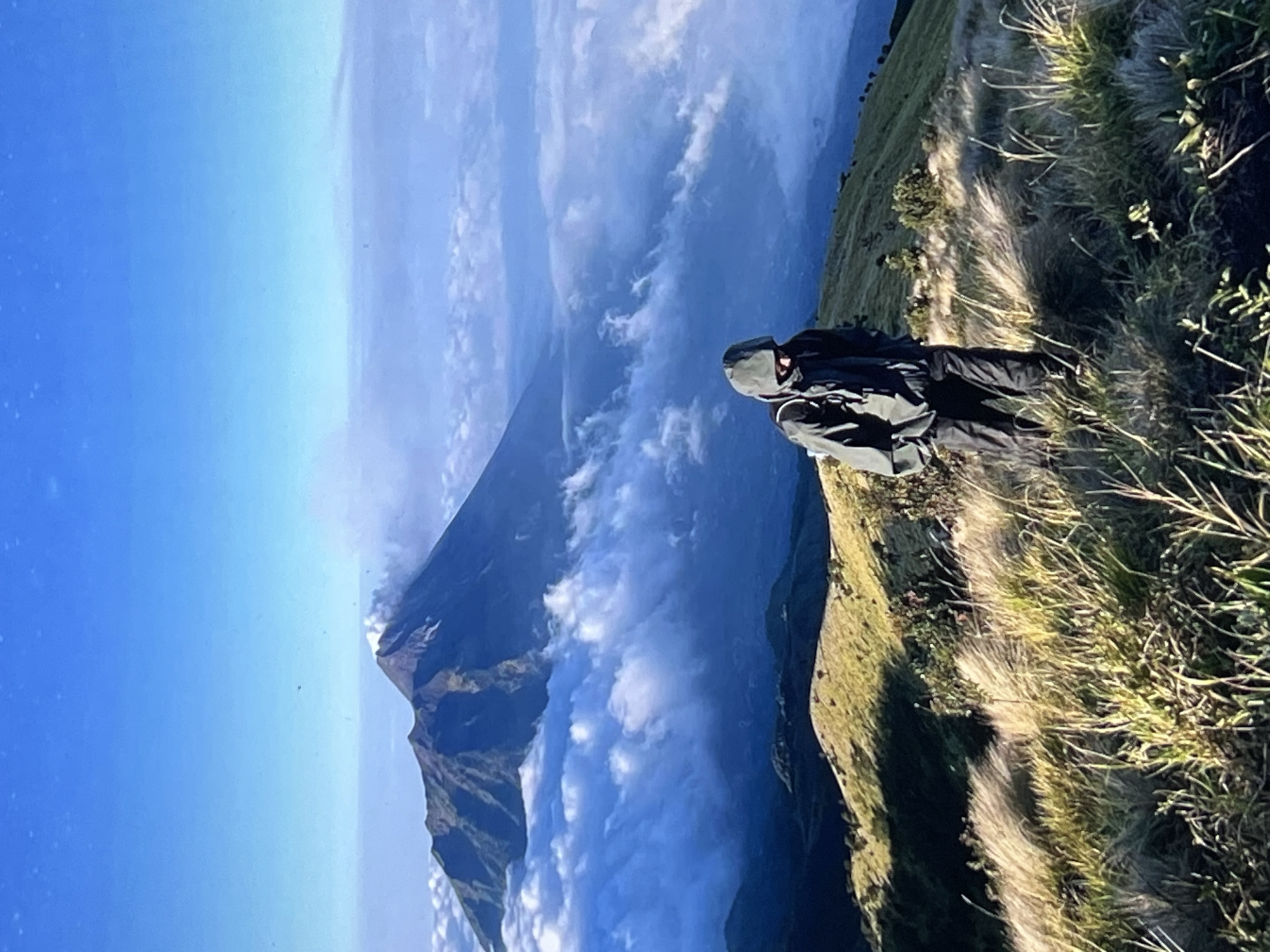A person in a dark jacket and trousers stands on a grassy, sloping hillside, looking out towards a large mountain peak that rises above a sea of clouds. The sky is a clear blue. The location is Unknown, Unknown.