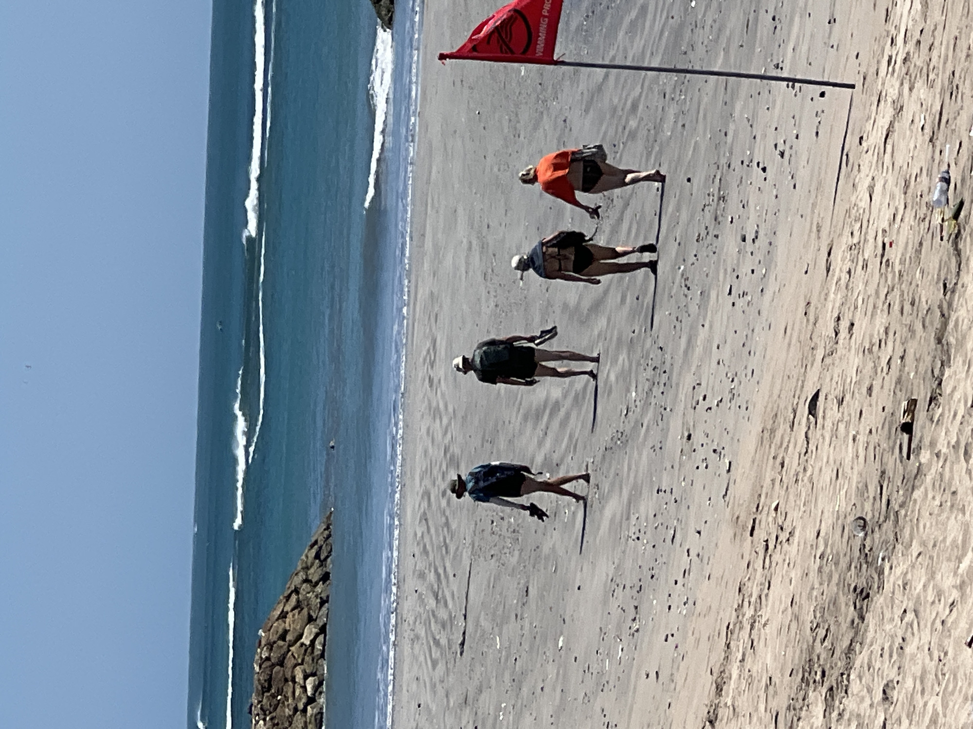 Four individuals walk along a sandy beach in Kuta, Indonesia, with the ocean and waves in the background. A red flag with a circular symbol and text is planted on the right side of the image. The sand is textured with ripples and scattered with small dark objects. In the distance, waves break on the shore under a clear blue sky. A rocky structure juts out into the water on the left side of the frame. Some litter is visible on the sand in the foreground.