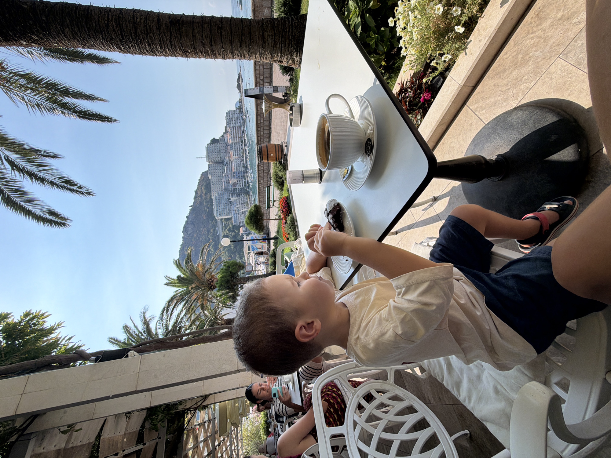 A young boy is seated at a table in an outdoor cafe, with a cup of coffee and a pastry in front of him. He is wearing a light-colored t-shirt and dark shorts. In the background, other patrons are visible, and beyond them, a body of water with boats and a city skyline can be seen. Palm trees frame the scene. The location appears to be the promenade in Bečići, Montenegro, with the city of Budva visible across the bay.