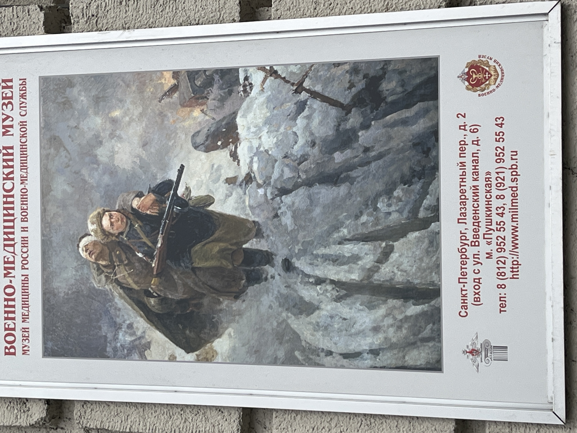 The media file shows a vertical outdoor sign for the Military Medical Museum, mounted on a textured, light-colored wall.
The left side of the sign features a painting depicting a harsh winter scene. Three figures, likely soldiers or military medical personnel, are shown in heavy winter clothing navigating a snow-covered, desolate landscape. One figure is carrying an injured comrade with a bandaged head, while another figure walks alongside, holding a rifle. The painting conveys struggle, determination, and care in a wartime or post-combat environment.
The right side of the sign presents text in Russian, providing details about the museum. The main title reads "ВОЕННО-МЕДИЦИНСКИЙ МУЗЕЙ" (Military Medical Museum), with the subtitle "МУЗЕЙ МЕДИЦИНЫ РОССИИ И ВОЕННО-МЕДИЦИНСКОЙ СЛУЖБЫ" (Museum of Medicine of Russia and Military Medical Service). The address is "Санкт-Петербург, Лазаретный пер., д. 2" (St. Petersburg, Lazaretny Lane, building 2), with an entrance specified as "(вход с ул. Введенский канал, д. 6)" (entrance from Vvedensky Canal street, building 6). The nearest metro station is "м. «Пушкинская»" (Metro "Pushkinskaya"). Contact information includes phone numbers "8 (812) 952 5543, 8 (921) 952 55 43" and the website "http://www.milmed.spb.ru". Two distinct emblems are visible: one at the top right, featuring a medical cross and serpents within a crest, and another at the bottom right, resembling a winged architectural column.