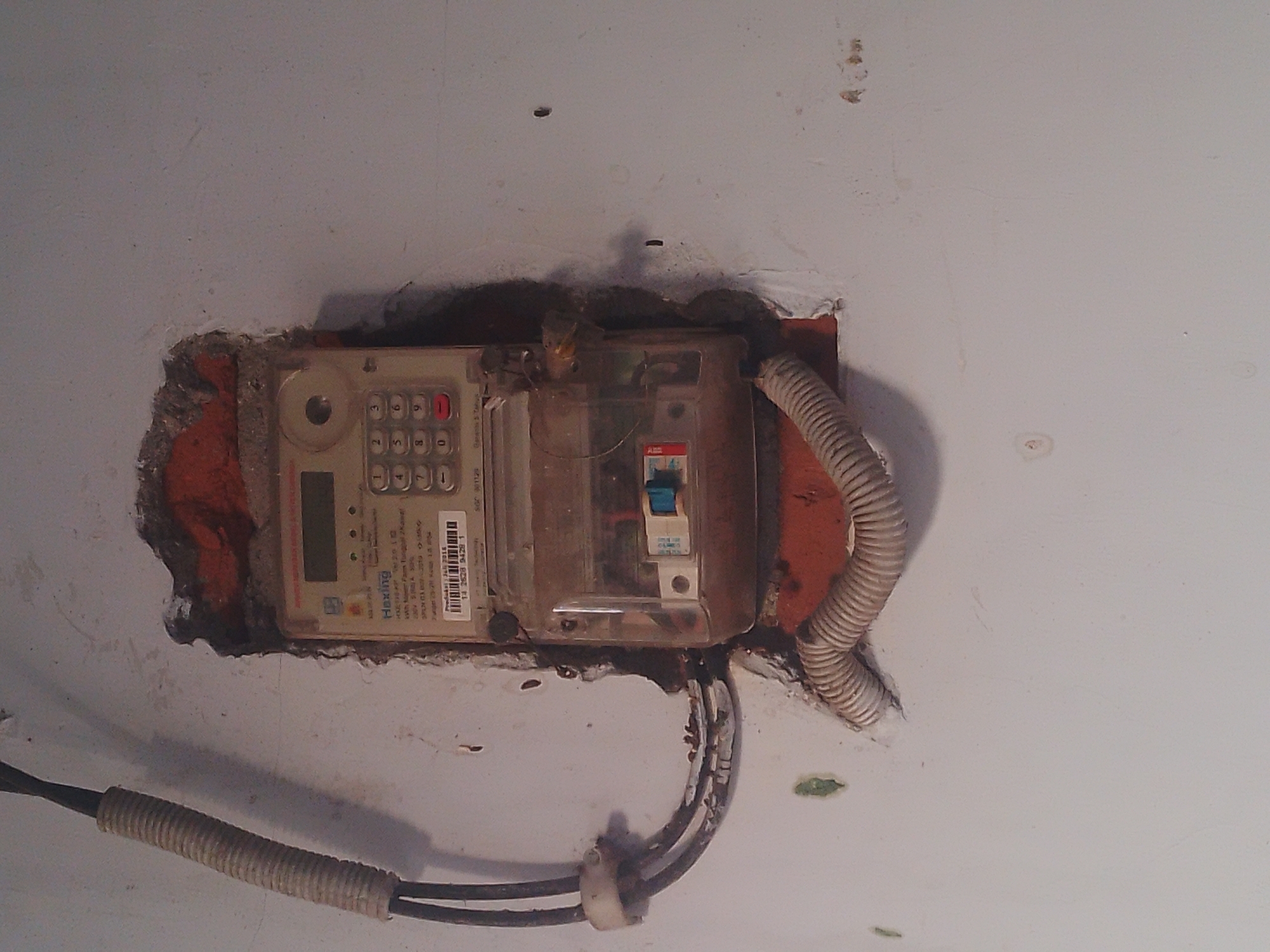 In the city of Pelem, Indonesia, a weathered, off-white wall serves as the backdrop for an electricity meter embedded within a rough, damaged opening. The meter, a Hexing model, features a digital display, a keypad with numerical buttons, and a clear plastic casing revealing internal components. Two thick black cables, covered by a corrugated plastic conduit, enter the meter from the left. A coiled, flexible conduit emerges from the bottom of the opening, disappearing downwards. The surrounding wall shows signs of age and wear, with cracks, chipped paint, and some green discoloration. The lighting appears to be ambient, casting subtle shadows.