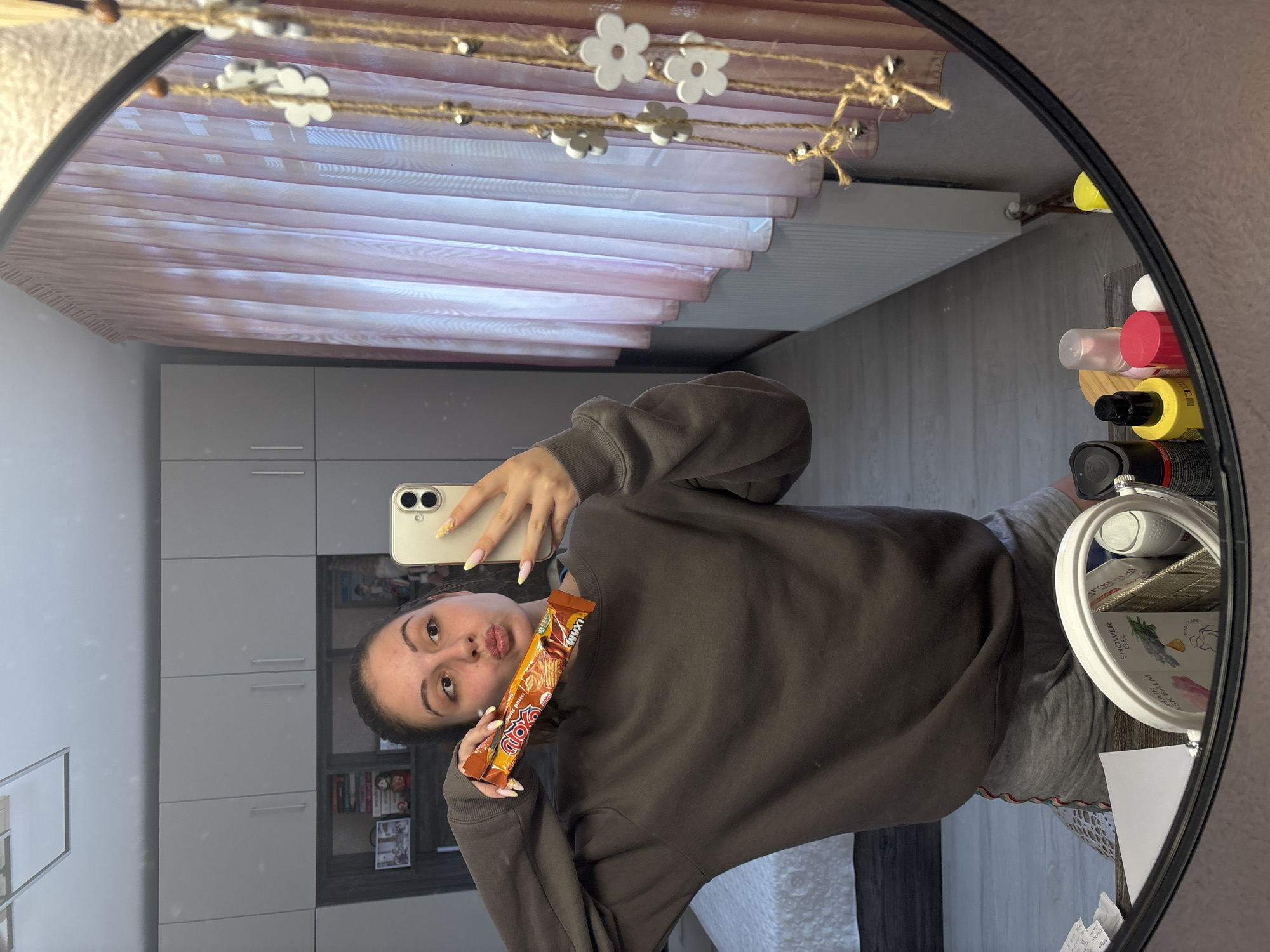 A young woman is reflected in a round mirror, taking a selfie. She holds a candy bar in one hand and a smartphone in the other, posing with a pouting expression. She wears a grey sweatshirt and sweatpants. The background shows a room with light grey cabinets and a striped pink and white curtain. Cosmetics and personal care items are visible on a shelf within the mirror's reflection. The location of the image is unknown and cannot be confirmed as Bulgarovo, Bulgaria.