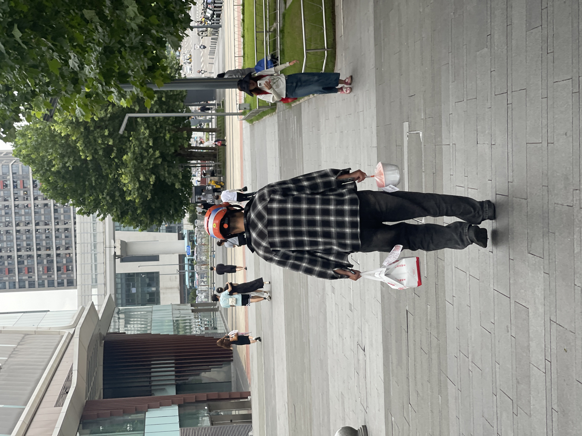 The image depicts a man walking up a set of stone stairs, carrying two bags. He is wearing a plaid shirt, dark pants, and a red and blue helmet. The man is walking away from the camera, towards a sidewalk lined with trees and buildings. In the background, there are several people walking on the sidewalk and a street with vehicles. The location is identified as the Chongqing High-tech Zone, China. The scene appears to be a public space, possibly a commercial or mixed-use area within the city. The lighting suggests it is daytime.