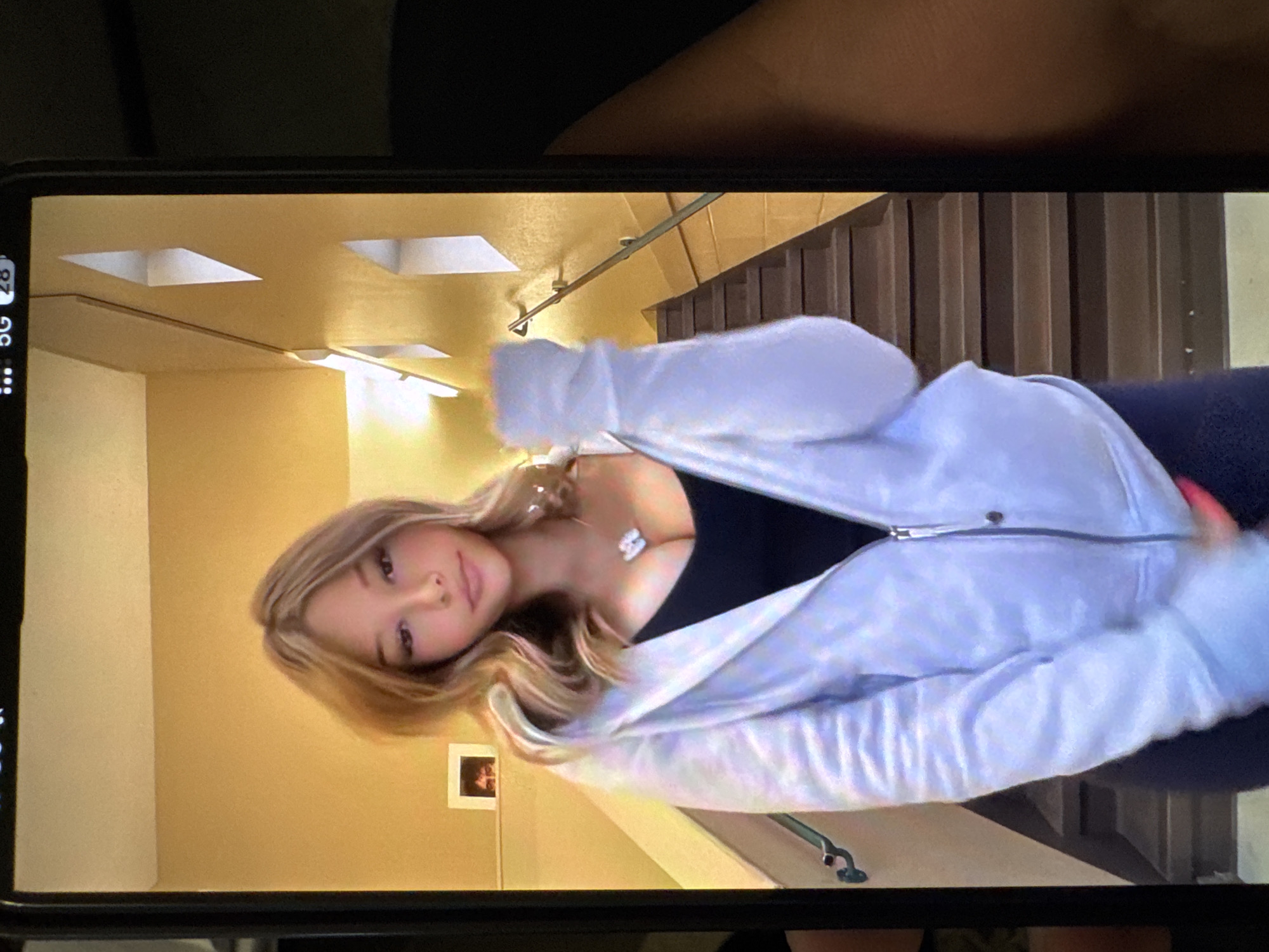 A person with blonde hair and a white jacket poses in front of a set of stairs. The background includes yellow walls, ceiling lights, and a framed picture on the wall. The image is displayed on a smartphone screen. There is nothing to suggest this image was taken in Yerevan, Armenia.