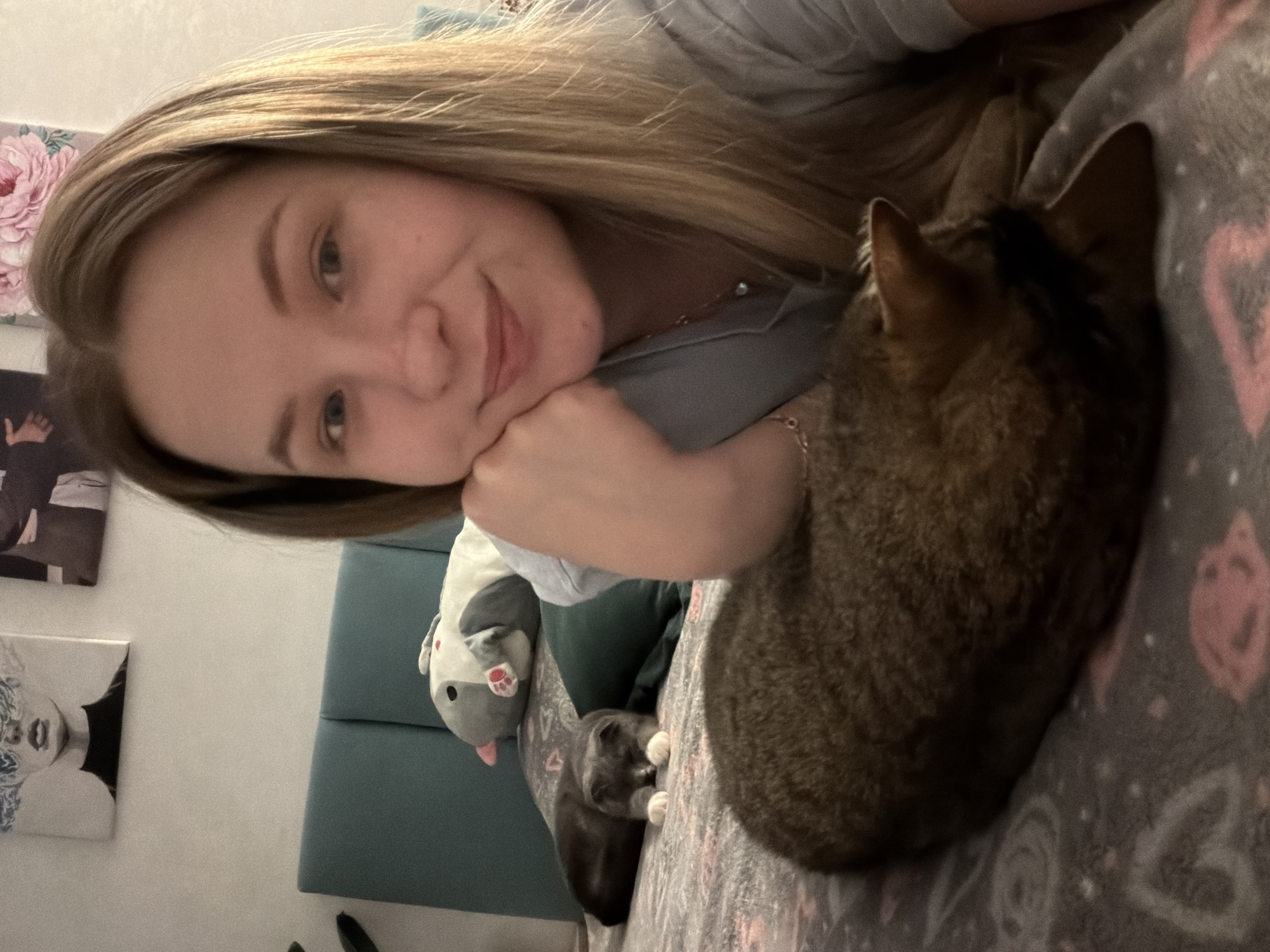 A young woman with long blonde hair is lying on a patterned bed, resting her face on her hand and smiling at the camera. A brown tabby cat is curled up next to her on the bed. Behind the woman is a blue headboard and a wall decorated with two pieces of artwork and a plush toy. Based on the objects present in the image, it is most likely taken in a private residence, but the location of the city of Chaikovsky, Russia cannot be determined from the image alone.