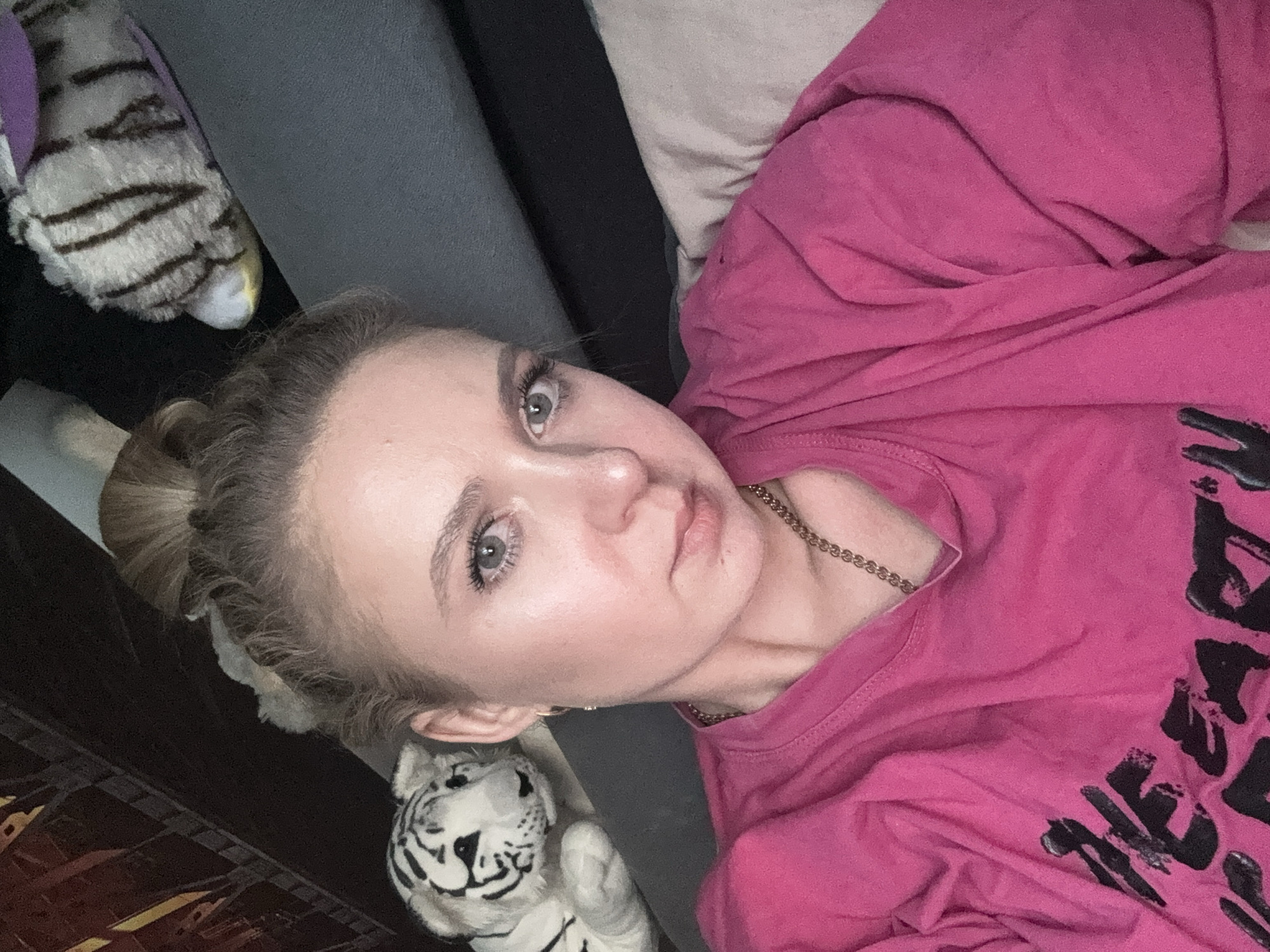 A young woman with blonde hair pulled up in a bun is captured in a close-up selfie. She is wearing a bright pink t-shirt with black text on it and a gold chain necklace. Her expression is neutral. To her left, a plush toy tiger is visible, and another plush toy, possibly a tiger as well, is seen in the background to the right. The setting appears to be indoors, possibly a living room, with a couch and a dark background suggesting furniture or a wall. The lighting is soft, highlighting the woman's facial features.