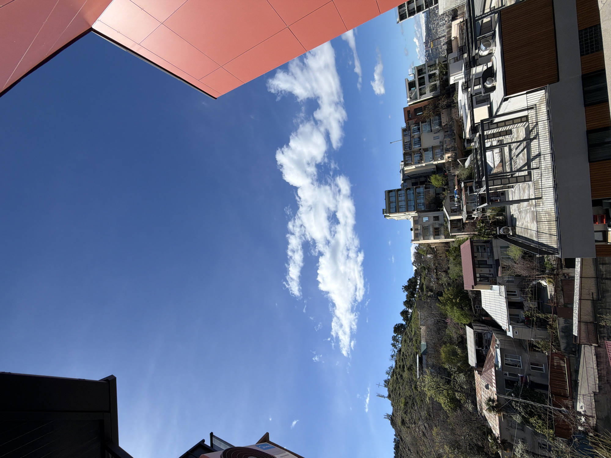 Captured in Tbilisi, Georgia, the image presents an upward-angled view showcasing a blend of urban and natural elements. In the sky, cumulus clouds dot a clear blue expanse, partially framed by the angular architecture of adjacent buildings, one featuring rust-colored paneling. A mix of mid-rise apartment buildings and private homes line the background, some nestled on a steep, wooded hillside. The scene is a juxtaposition of modern construction against the organic texture of the city's natural landscape.