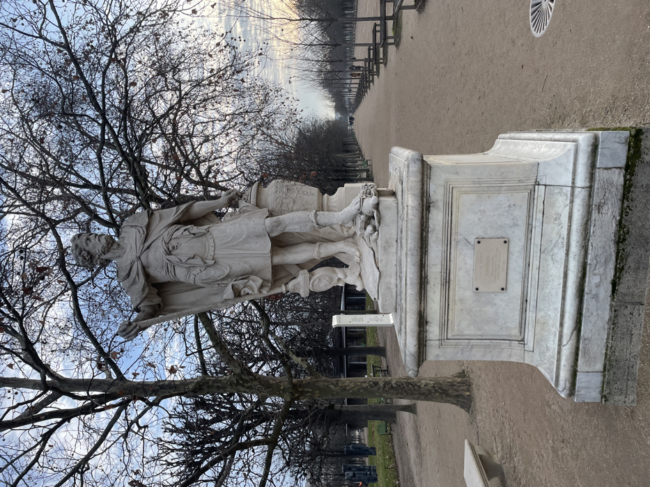 The image displays a white marble classical statue of a bearded male figure, centrally located on a wide gravel path within the Tuileries Garden, Paris, France. The figure stands on a multi-tiered marble pedestal, draped in a toga-like garment, holding a staff vertically in his raised left hand and an ornate, lidded urn in his right hand. A rectangular plaque is affixed to the front face of the pedestal.
The scene is set under an overcast sky, with numerous deciduous trees bearing bare branches flanking the path. The wide, light brown gravel path extends into the deep background, lined with benches on the right side. To the left of the statue, a grassy area precedes further bare trees and a distant building with large windows, along with other indistinct sculptural forms. Several indistinct human figures are visible walking in the far distance along the main avenue. The environment is a well-maintained public park during a cool season.