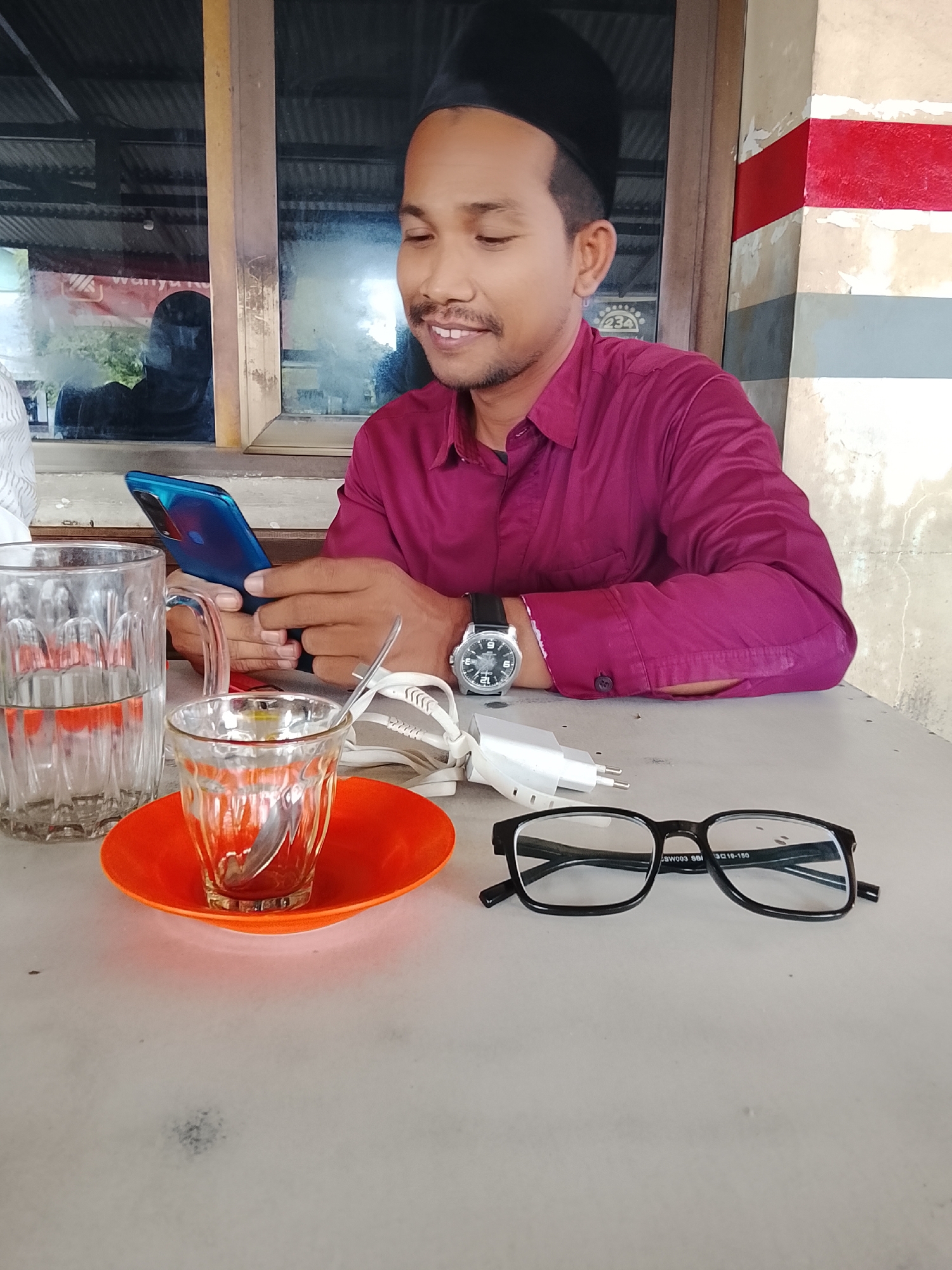 A man wearing a dark peci and a maroon collared shirt is seated at a table, holding a blue smartphone in his left hand. On the table in front of him are several items: a clear glass of water with a textured pattern, a small clear glass with a spoon on an orange saucer, a white electrical adapter, and a pair of black-rimmed glasses. The man is looking down at his phone with a slight smile. In the background, a window reveals a reflection of another person wearing dark clothing. The setting appears to be an outdoor or semi-outdoor space, possibly a warung or a casual eatery, with a red and white striped pillar visible to the right. The table surface is a light-colored, possibly concrete or stone material. The overall lighting suggests it is daytime. The man's watch on his left wrist is a silver analog watch with a black strap. The inscription on the glasses appears to read "SW003 SBI 3016-150". Based on the architecture and the presence of the red and white stripes which are common in Indonesian public spaces and local shops, this scene is consistent with a location in Lhokseumawe, Indonesia.