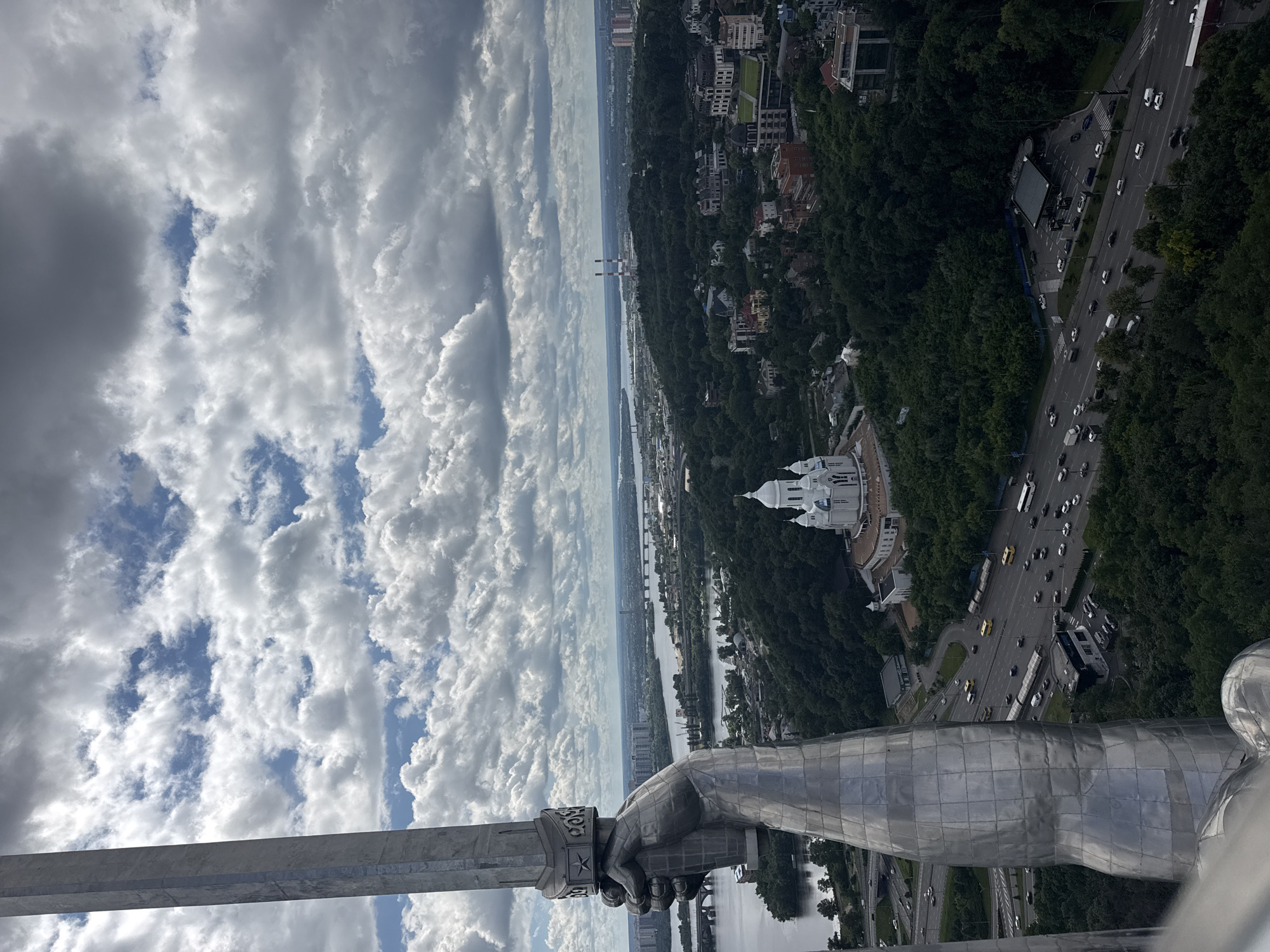 This is a medium-angle aerial photograph taken from a high vantage point overlooking a section of Kyiv, Ukraine. In the foreground, a large, metallic sculpture of a hand holding a sword dominates the left side of the frame, its polished surface reflecting the sky. Beyond the sculpture, the Dnieper River flows across the middle ground, spanned by several bridges. On the opposite bank, a lush green hillside is dotted with buildings, including a prominent white church with blue domes, and a cluster of modern residential buildings and industrial structures, including two tall smokestacks with red and white bands. A highway with moderate vehicle traffic runs along the base of the hill. The sky is filled with dramatic, heavy white and gray clouds.