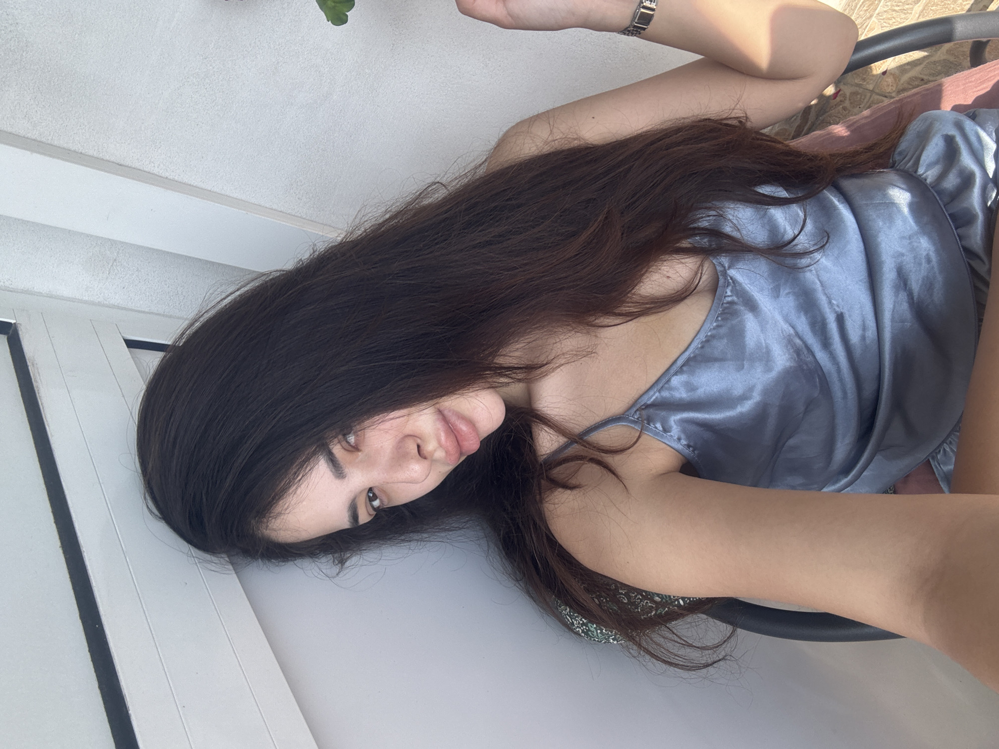 A young woman is captured in a close-up, angled shot, lying on her side and looking towards the camera. She has long, dark brown hair that falls over her shoulders and arm. Her face, framed by her hair, features full lips slightly parted and dark eyes with a calm expression. She is wearing a light blue or silvery-grey satin-like camisole or top with thin straps. A silver-colored watch is visible on her left wrist.
The background shows a white, textured wall and white structural beams or trim, suggesting an indoor or covered outdoor setting like a balcony or patio. A dark vertical line, possibly a window or door frame, is present on the left. Faint hints of a light brown, possibly tiled, floor can be seen, along with parts of a dark chair or furniture behind her. A small green plant is partially visible in the upper right. The lighting suggests bright daylight. There is no visible text in the image.