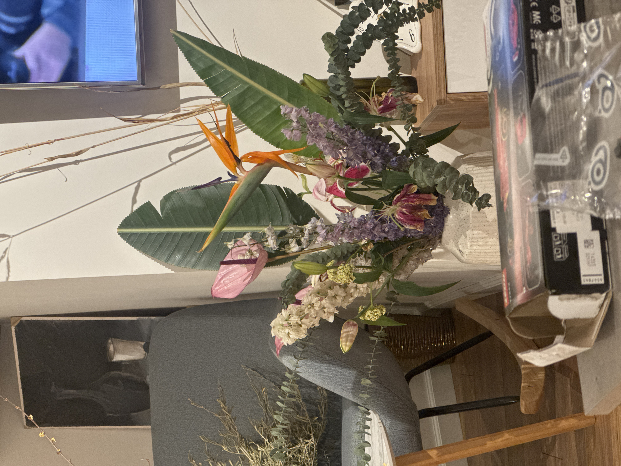 A medium, eye-level shot depicts an interior featuring a floral arrangement. In the foreground is a bouquet that includes bird of paradise flowers, other assorted blossoms, and eucalyptus branches. The arrangement sits on a table, adjacent to a box or package with visible printed text and designs.
In the background, partially obscured by the bouquet, there's a gray upholstered object, possibly a chair or sofa. Behind that, there's a framed print or photograph visible on the wall. The room appears to be well-lit, and the scene suggests a domestic setting.