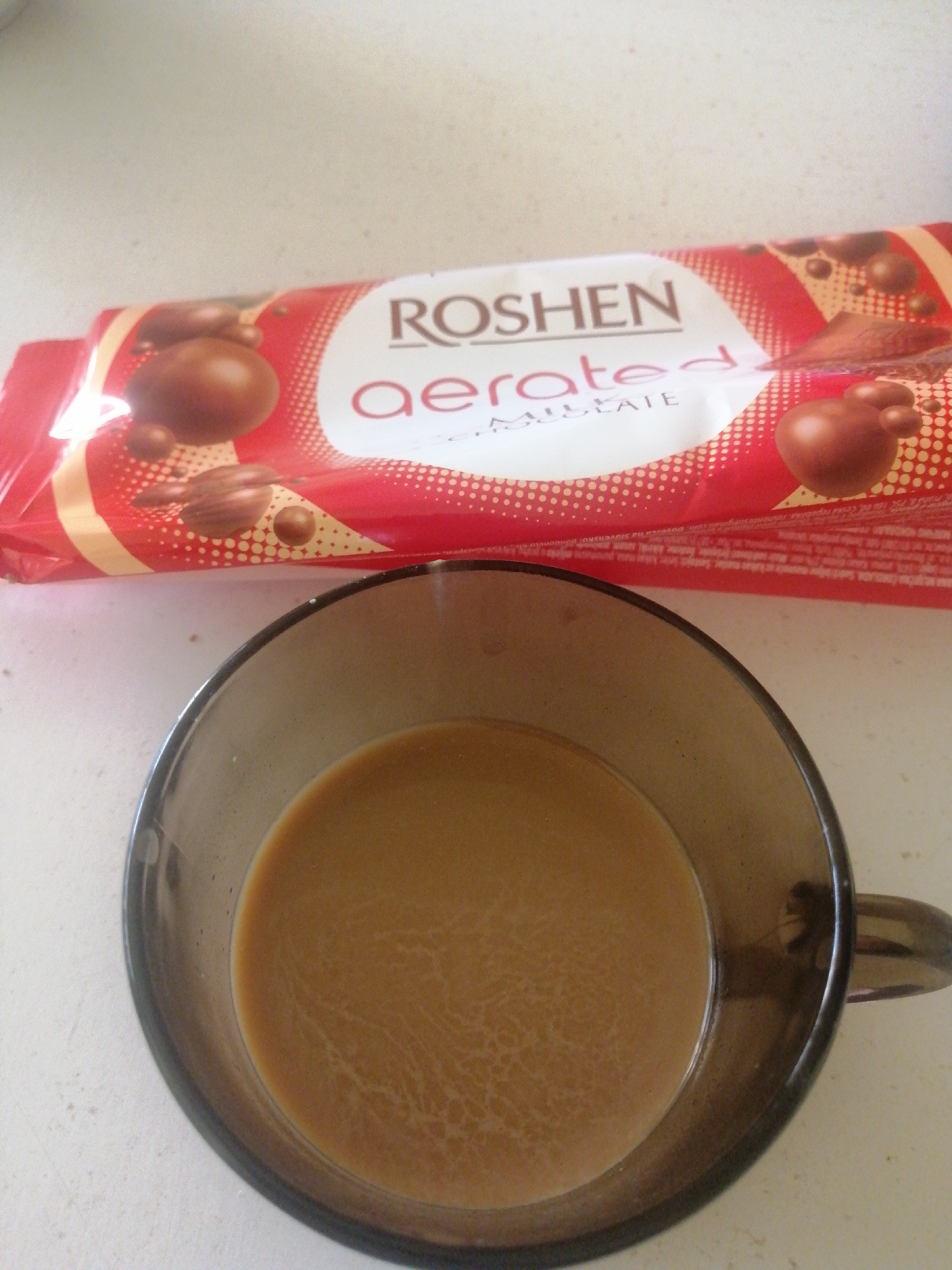 A top-down, eye-level shot shows a brown coffee mug containing coffee with creamer in front of a wrapped Roshen Aerated Chocolate candy bar. The background is a white surface. There are no people visible. The location cannot be determined as Sofia, Bulgaria, from the image alone.