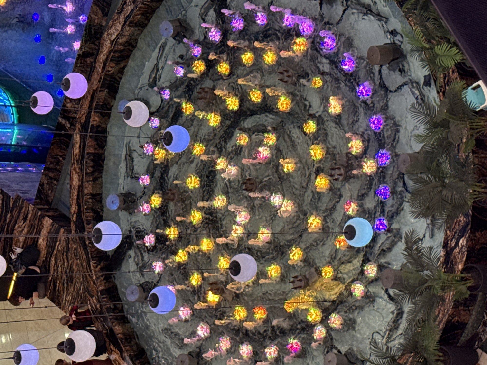 The image shows an indoor fountain with colorful lights. Several people are visible around the edge of the fountain, with one person in the foreground appearing to be sitting on the edge. The fountain itself has multiple water jets that are illuminated with various colors, including blue, purple, yellow, and pink. Hanging from above are several spherical lights, some of which are also illuminated. Plants in pots are visible in the lower corners of the image. The fountain is surrounded by a decorative stone-like material. The overall impression is of a brightly lit, decorative indoor space, possibly a shopping mall or public area.