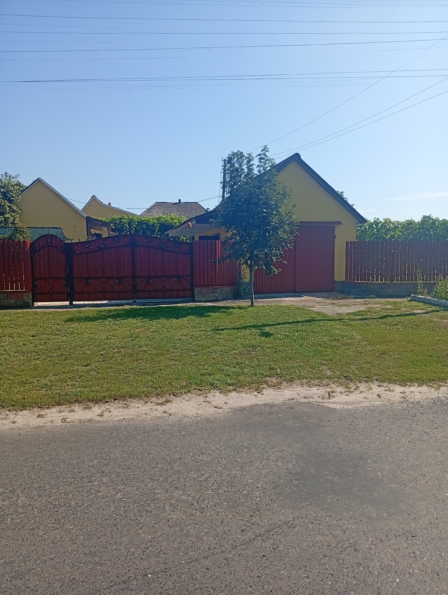 The image depicts a residential area on a clear day, likely in Khomutets, Ukraine, as indicated by metadata. The scene features a property enclosed by a red wooden fence with decorative metalwork on the gate. Behind the fence, a yellow-painted house or garage with a red door is visible, flanked by more fencing and greenery. A young tree stands in the foreground on the property. A paved road runs along the bottom of the image, with a strip of grass separating it from the fence. Power lines are strung across the clear blue sky. No people or animals are present in the frame. The overall impression is one of a quiet, sunlit rural or suburban environment.