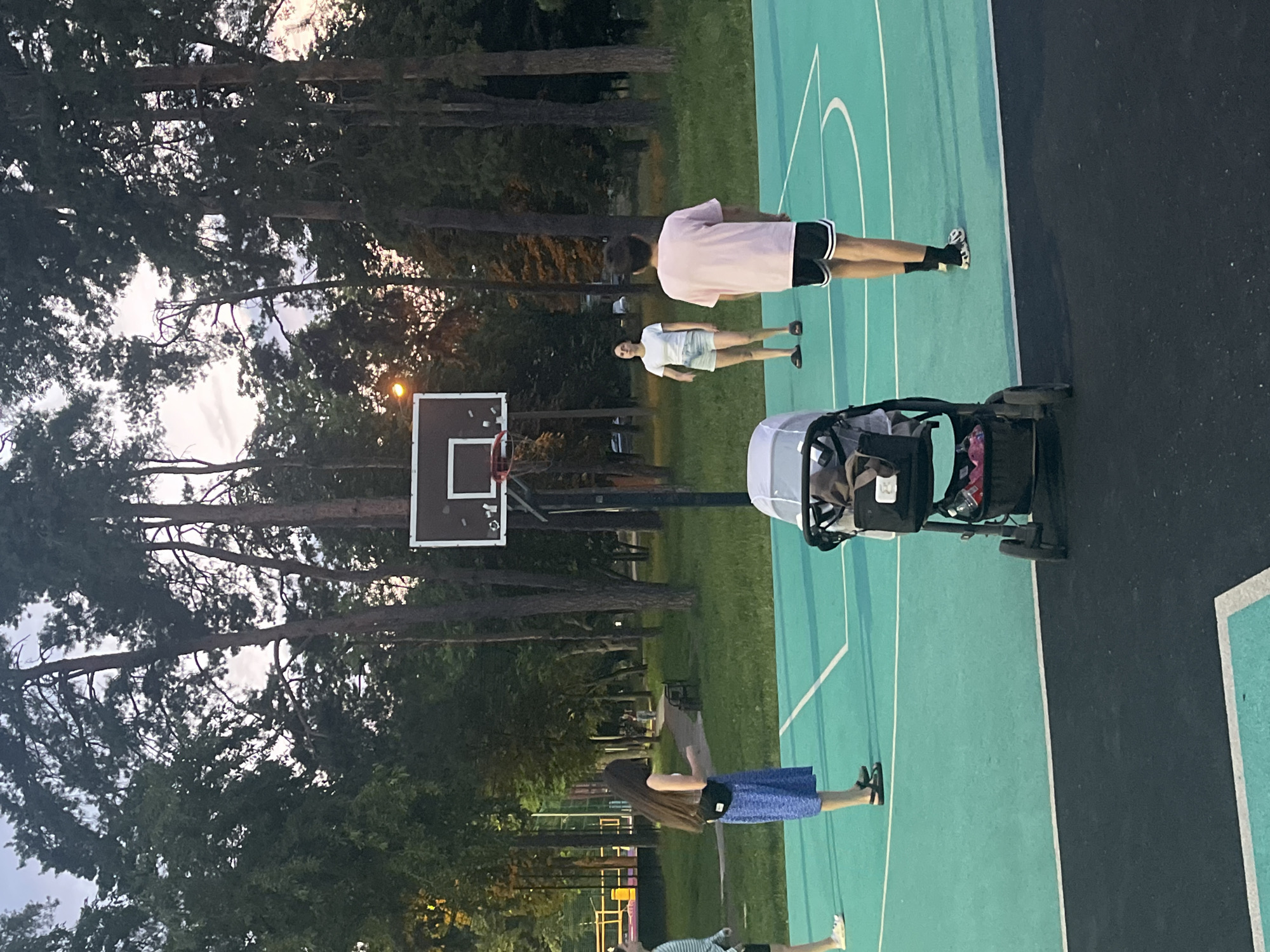 The image shows an outdoor basketball court in a park-like setting, likely in the city of Krasnodar, Russia, given the context. A turquoise-colored court with white markings is visible, bordered by a dark asphalt area. Several people are present on the court. In the foreground, a grey stroller is parked. To the left, a person wearing a blue skirt and a fanny pack stands facing away from the camera, seemingly interacting with someone out of frame to the left. Further back, on the court, two individuals are standing: one in a white t-shirt and denim shorts, and another in a pink t-shirt and black shorts with white stripes. A basketball hoop with a backboard is positioned behind them. The background features tall trees with lush green foliage and a dusky sky, suggesting it is late afternoon or early evening. The overall scene depicts a casual recreational activity taking place in a public park.