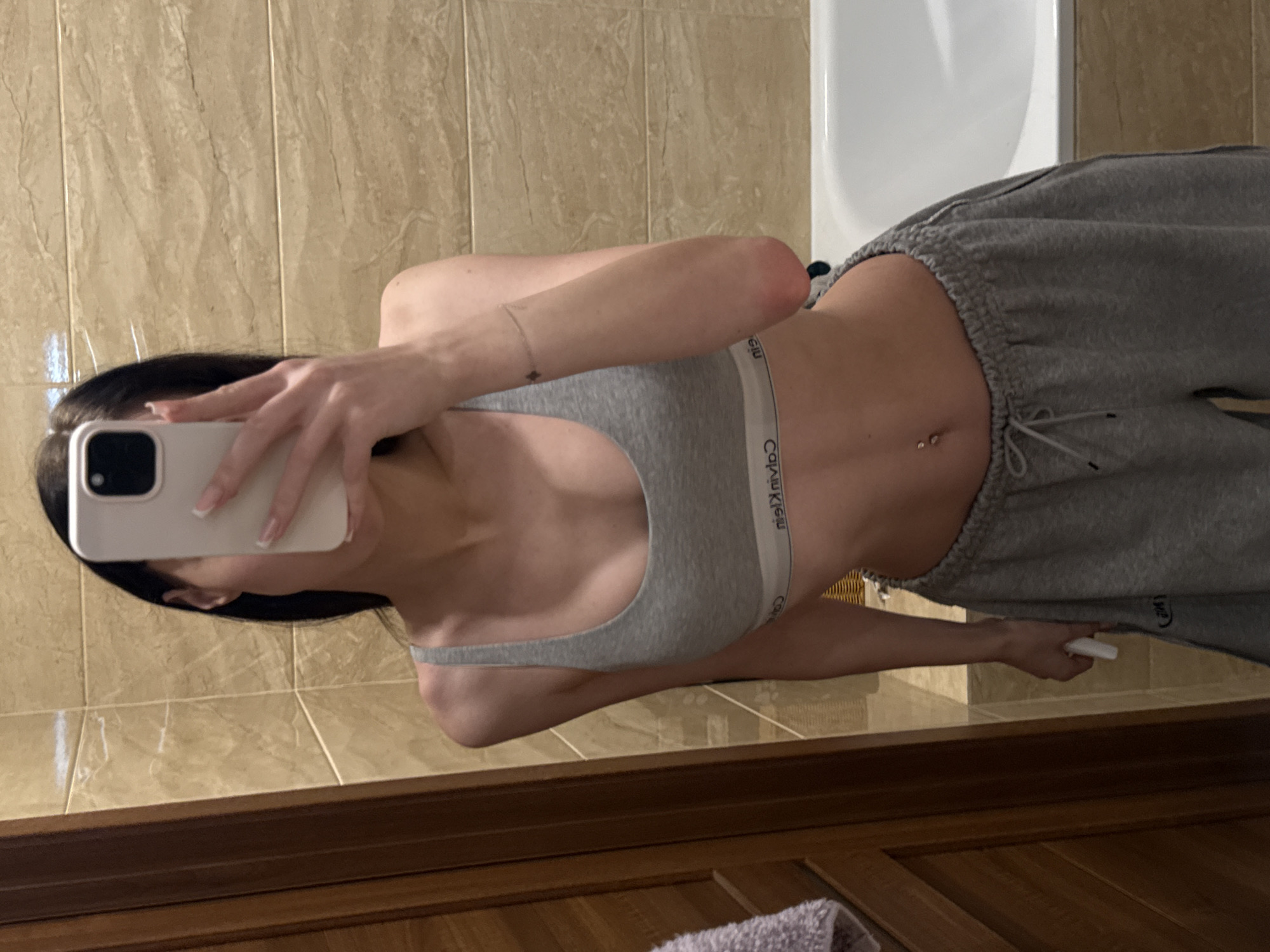 The image is a vertical, medium shot of a young woman taking a mirror selfie in a bathroom. The woman has long, dark hair and is wearing a grey sports bra and grey sweatpants. She is holding a white smartphone in her right hand, angled to capture her reflection. A small, diamond-shaped charm is visible on a bracelet on her left wrist. Her navel is pierced with two small studs.
The bathroom features light brown, marbled tiles on the walls and a white bathtub is partially visible in the background. The flooring is not fully visible, but wooden cabinets or furniture are present at the bottom of the frame. The lighting suggests it is indoors, likely in the evening or a room with artificial light.
The woman's expression is not clearly visible due to the angle and the phone obscuring her face. The overall mood is casual and personal, typical of a selfie taken in a private space. The text "Calvin Klein" is visible on the waistband of the sports bra. The image is set in Khimki, Russia.