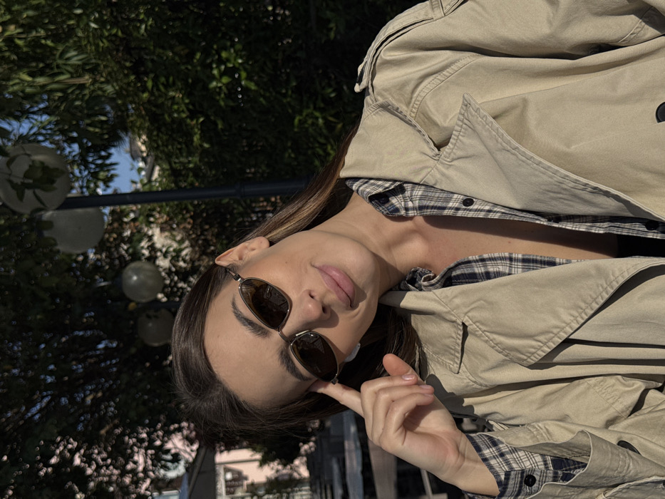 An adult female is positioned outdoors in Sirmione, Italy, illuminated by direct sunlight. She possesses long, dark brown hair and wears small, dark-tinted, oval-shaped sunglasses. Her attire consists of an open beige trench coat layered over an unbuttoned blue and white plaid shirt, revealing a dark undergarment. Her right hand is raised, with fingers resting on her right temple near the frame of her sunglasses. She exhibits a subtle smile. The background is composed of dense green foliage, likely from trees or bushes, with several spherical white outdoor lamps mounted on a dark pole visible within the greenery. A light-colored building is partially discernible through the leaves on the left, and glimpses of clear blue sky appear above the foliage.