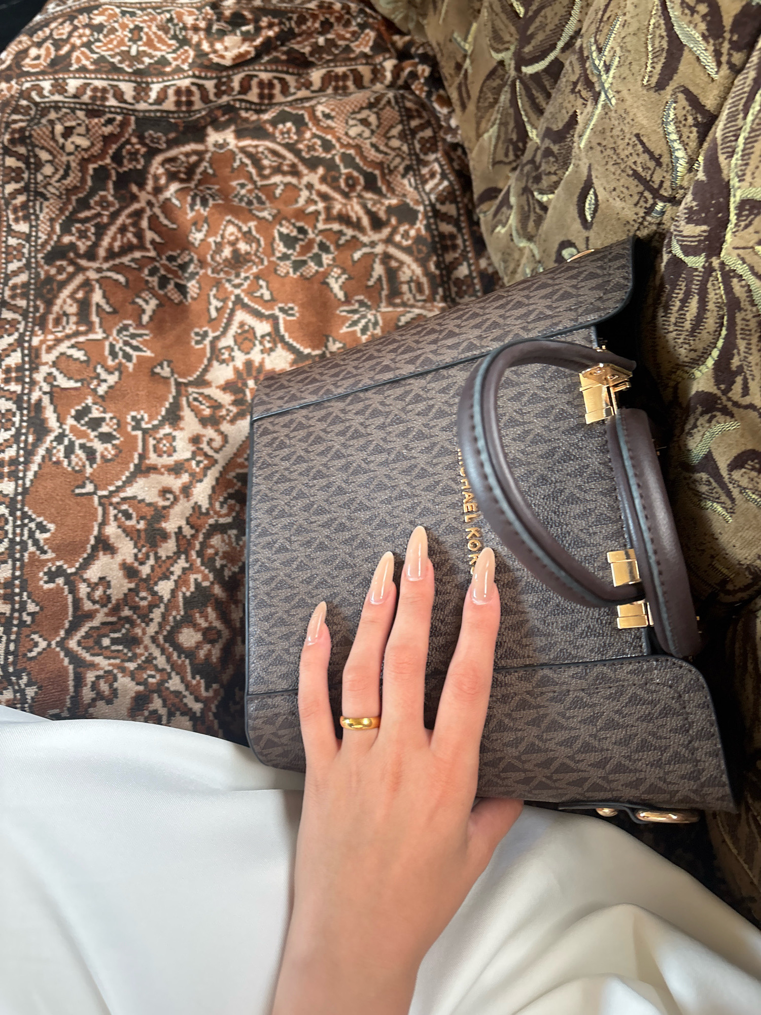 A person's left hand, adorned with long, light-colored artificial nails and a gold ring on the ring finger, rests on a brown and tan patterned fabric bag. The bag has a repeating geometric pattern in a darker shade of brown and features a dark brown leather handle with gold hardware. The bag is placed on a brown and cream patterned rug. To the right of the bag is a section of a brown and gold patterned sofa. In the lower left of the image, a fold of white fabric is visible. The scene appears to be indoors. The image does not contain any information about the city of Ijevan, Armenia.