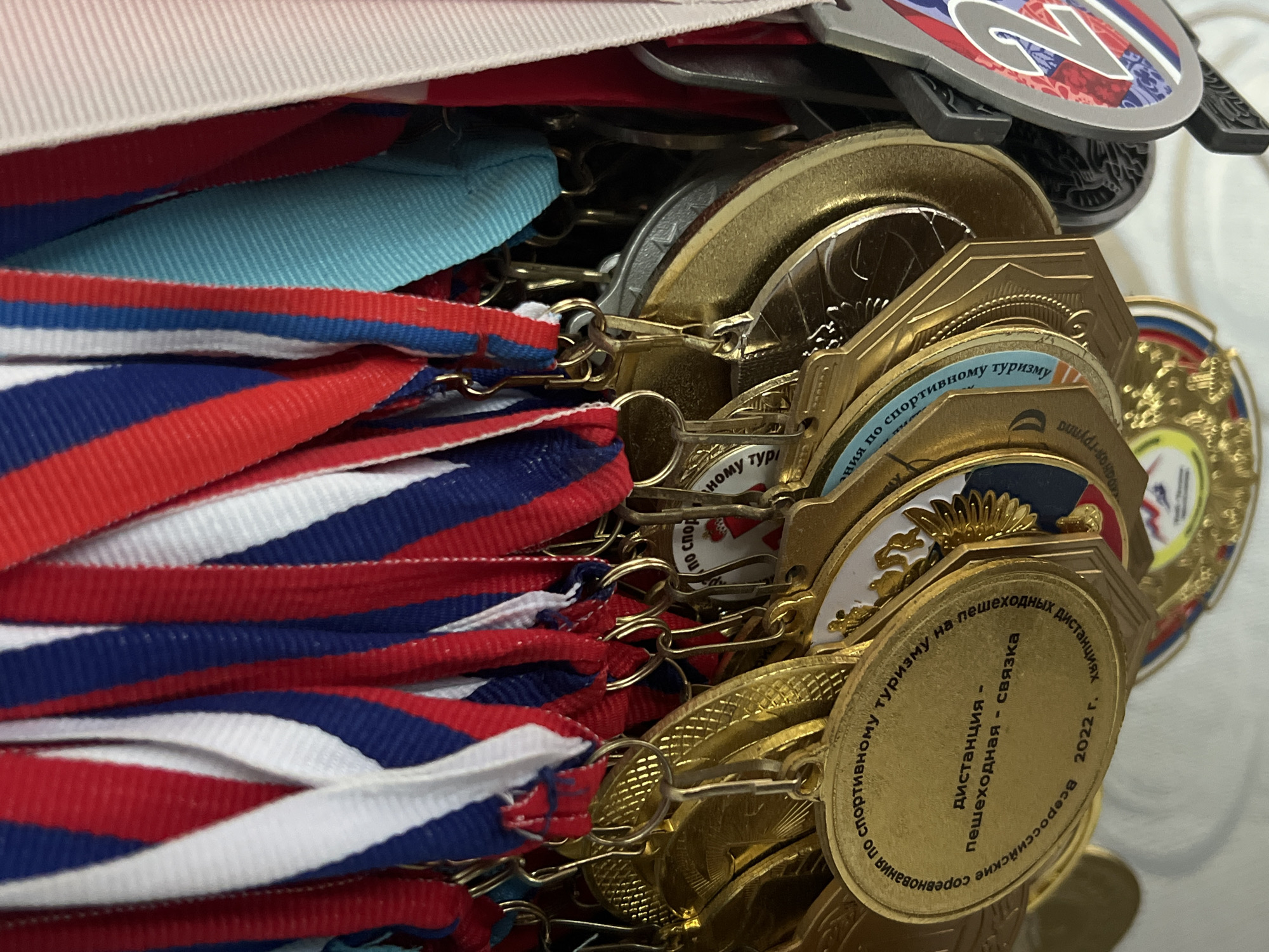 The image presents a close-up view of numerous sports medals hanging against a plain, light-colored background, located in the city of Chaikovsky, Russia. The medals are suspended by their ribbons from small, metallic hooks that are attached to the upper edge of each medal. The majority of the ribbons are vertically striped in red, white, and blue, indicative of the Russian national colors, while some are solid blue and one is light blue or turquoise. All visible ribbons display a finely ribbed texture.
The medals themselves are predominantly gold-colored, with some appearing silver. They vary in shape, including circular and shield-like designs. A prominent gold medal in the lower-left foreground features Cyrillic inscriptions: the outer circumference reads "Всероссийские соревнования по спортивному туризму на пешеходных дистанциях" (All-Russian competitions in sports tourism on pedestrian distances), while the central text states "дистанция - пешеходная - связка" (distance - pedestrian - team/bundle), with the year "2022 г." (2022 year) inscribed at the bottom. Another gold medal to its right partially displays an emblem resembling a double-headed eagle. On the far right, a silver-colored medal is prominently marked with the number "2" and incorporates red, white, and blue design elements, signifying a second-place achievement. No individuals are present in the frame.
