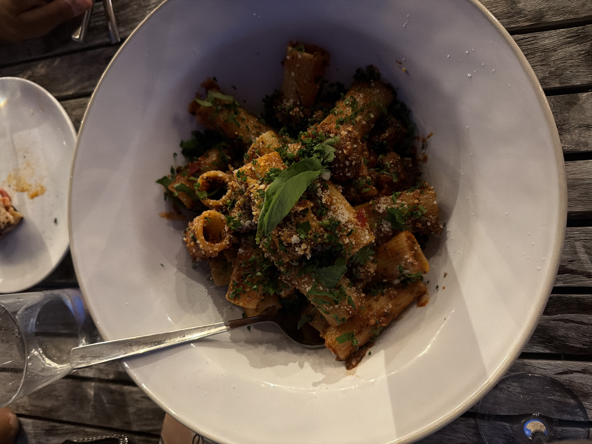 A top-down, close-up shot shows a white, shallow bowl filled with rigatoni pasta. The pasta is coated in a reddish-brown sauce and sprinkled with finely chopped green herbs and grated cheese. A large, fresh basil leaf garnishes the top of the pasta. A silver spoon rests on the left side of the bowl, with its handle extending out of the frame. The bowl sits on a dark wooden table. In the background, a glass of water is visible, and a portion of another plate with some food residue is also present. The scene appears to be set in Montauk, United States of America.