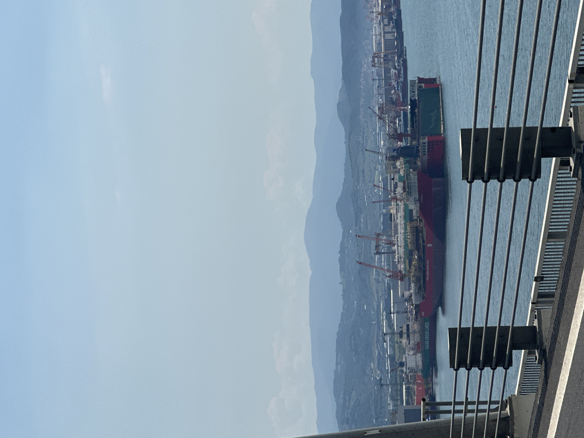 The image shows a view from a bridge, possibly a suspension bridge, looking towards a coastal industrial area. A large cargo ship is docked in the water, with cranes and other industrial structures visible along the shoreline. In the background, there are mountains. The city of Altınova, Turkey is likely the location. The bridge structure itself is visible in the foreground, including cables and supports.