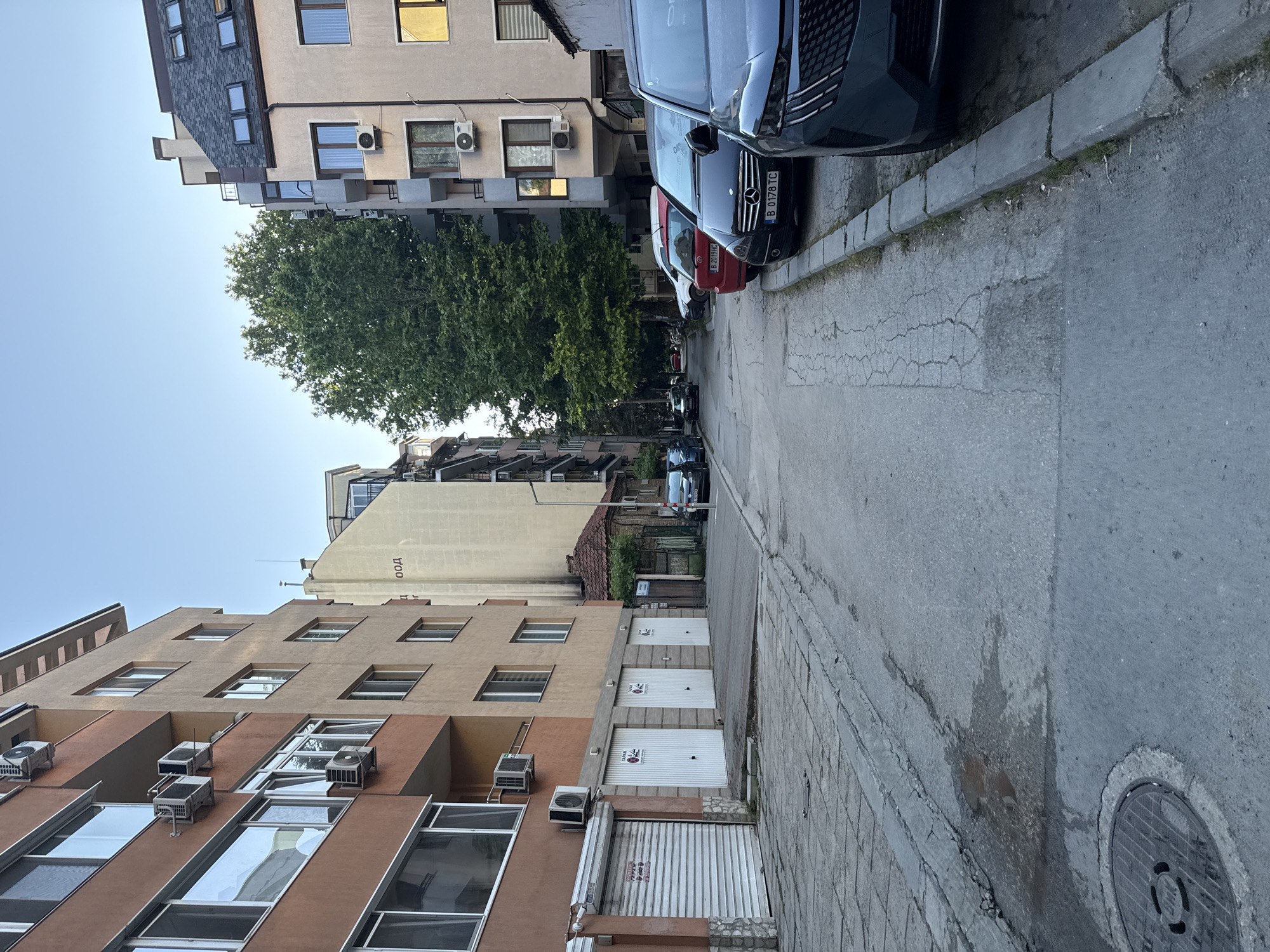 The image depicts a street scene in Varna, Bulgaria. On the left are multi-story apartment buildings with balconies and air conditioning units. The ground floor features several garage doors, some with signs. A sidewalk runs parallel to the buildings, featuring a manhole cover and patches of wet pavement. The street itself is asphalt, showing cracks and wear.
On the right side of the street, several cars are parked along the curb. These include a dark-colored SUV in the foreground, a black Mercedes, a red hatchback, and other vehicles further down the street. Behind the parked cars, another apartment building is visible, with a mansard roof and dormer windows. A large, leafy tree stands between the buildings, providing a significant amount of shade. The sky is clear and blue, suggesting it is daytime. The overall impression is of a typical urban residential area.
