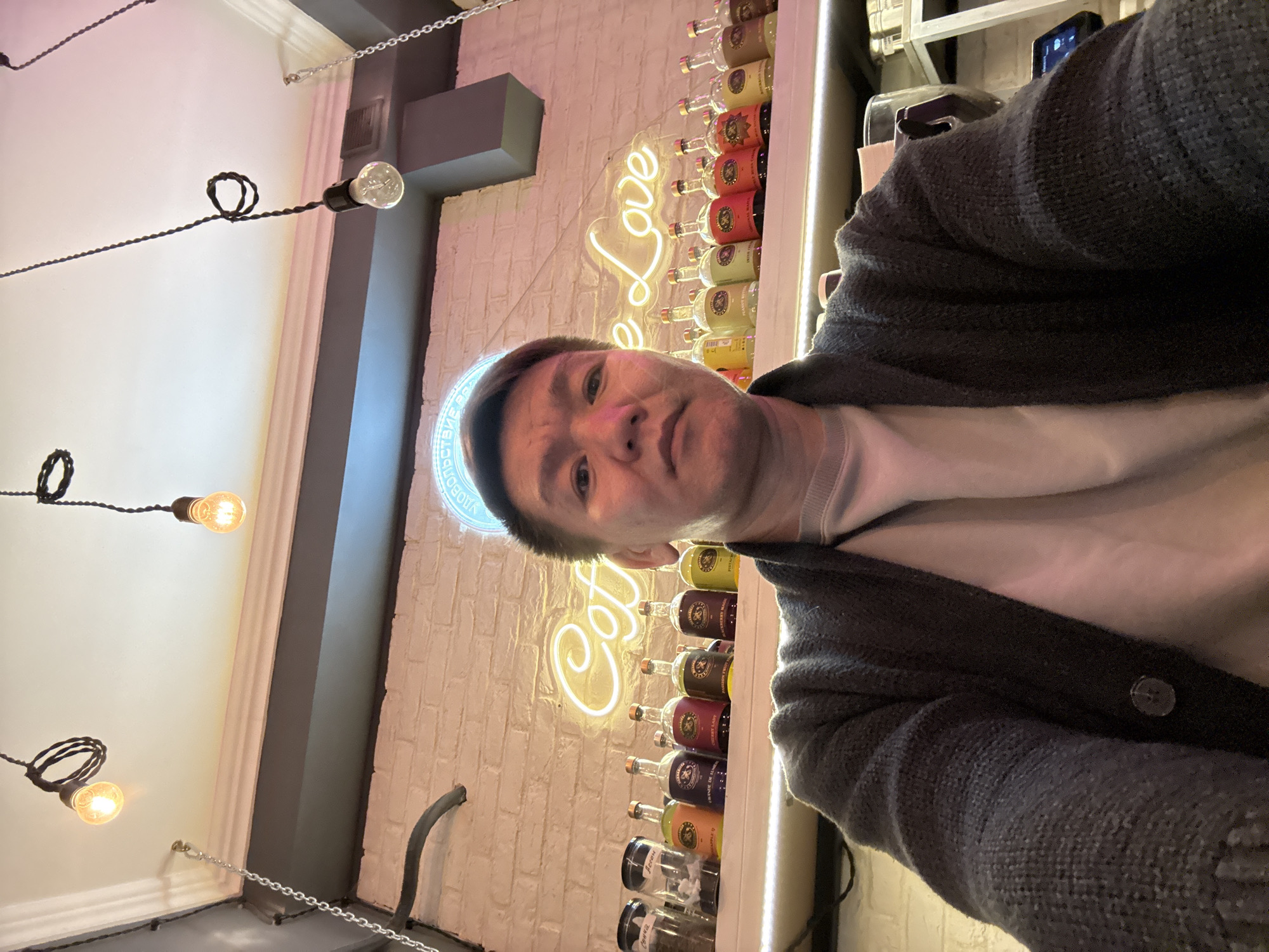 A medium, angled selfie shows a person with short brown hair and a light complexion, wearing a light-colored shirt and a dark gray cardigan, standing inside a space with a neon sign that reads "Coffee Love." Behind them, a white brick wall displays a shelf with multiple bottles and jars, likely liquor. Exposed light bulbs hang from the ceiling on twisted cords. The location is in Khimki, Russia, as part of the neon sign indicates "В Химках" which means "In Khimki" in Russian.