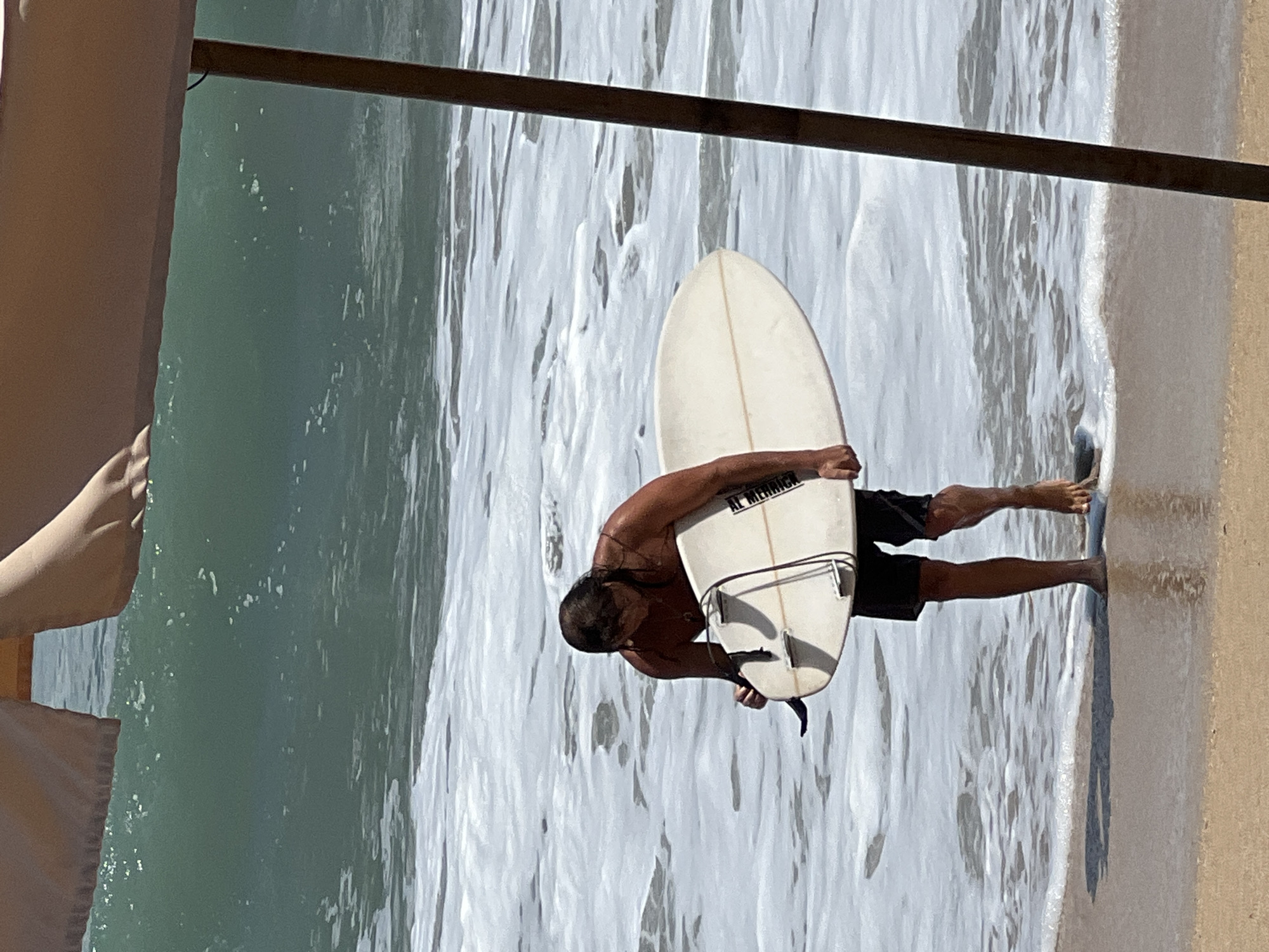 A person is standing in the ocean at the beach in Kuta, Indonesia, holding a surfboard. The person is wearing black shorts and has long, dark hair. The surfboard is white with fins and a leash. The ocean is calm with small waves and white foam. The beach is sandy and there is a wooden pole in the foreground. The sky is not visible. The overall scene depicts a surfer preparing to enter or exit the water in a tropical location.