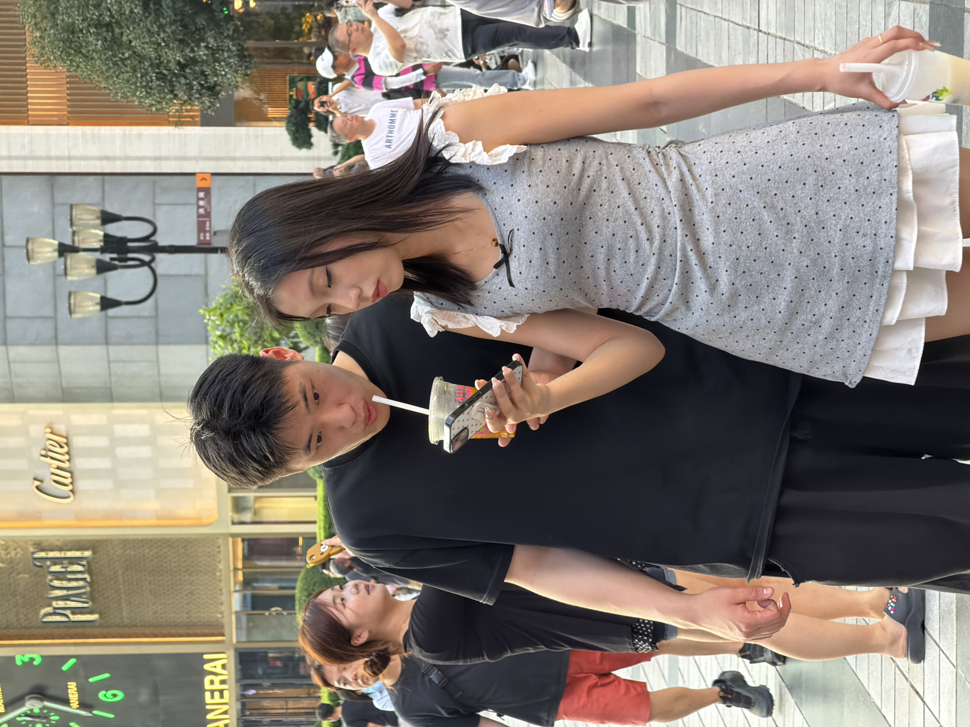 In a city scene at Yuzhong District, China, a young man in a black t-shirt and black shorts is observed holding a cup with a straw to his mouth, while simultaneously looking at a smartphone. A young woman stands beside him, also looking at the same smartphone held by the man, with her left hand near his. She is wearing a grey polka-dot dress with ruffled white trim at the sleeves and a white underskirt. In the background, other people are visible walking, with some holding up phones, possibly taking pictures. Storefronts with prominent signage for "Piaget" and "Cartier" are situated behind them, suggesting a shopping or commercial area. The environment appears to be outdoors, on a paved walkway, with some greenery and lampposts also present.