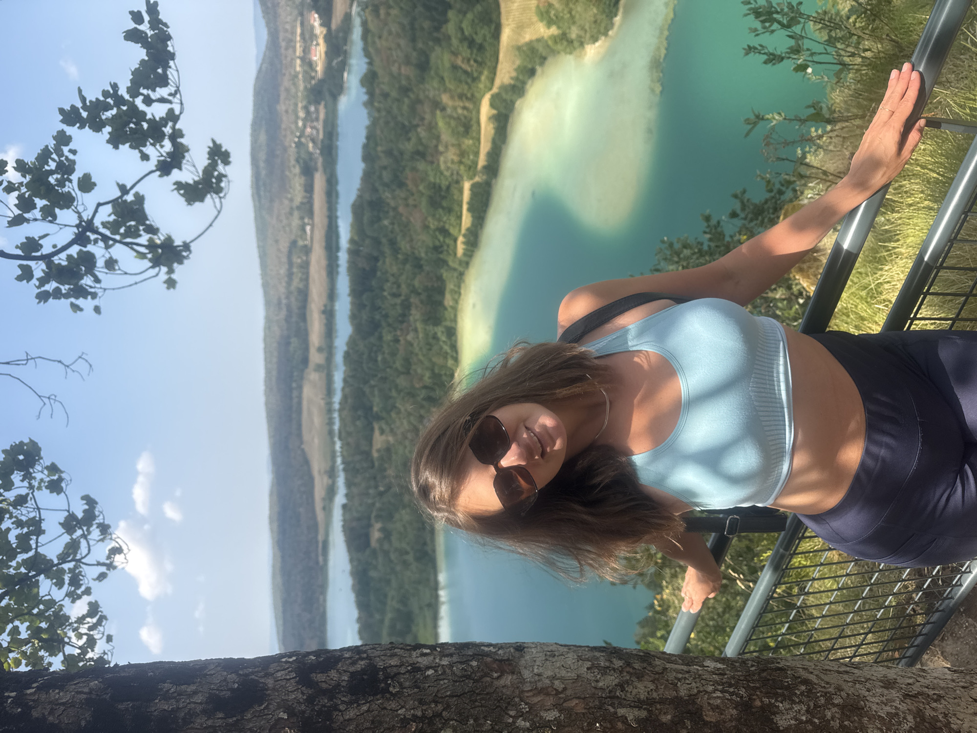 A woman with brown hair and sunglasses is standing on a railing next to a tree. She is wearing a light blue sports bra and dark blue leggings. Behind her, there are several bodies of water and a forested landscape under a clear blue sky. The scene appears to be a scenic viewpoint overlooking lakes and hills in the commune of La Chaux-du-Dombief, France.