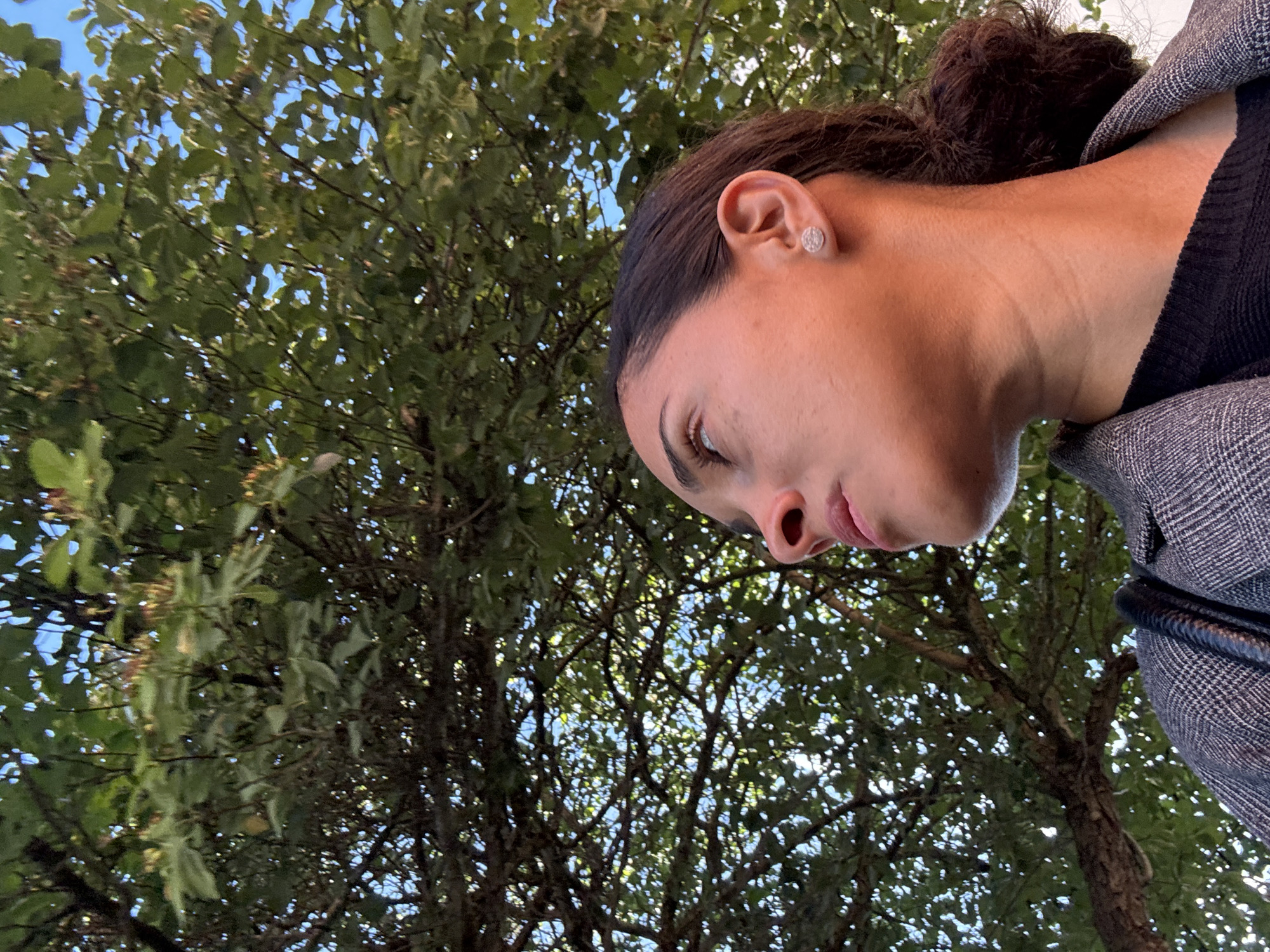 The image is a medium-shot, low-angle photograph of a woman looking upwards against a backdrop of green tree foliage and a blue sky. The woman has brown hair pulled back into a ponytail and is wearing a grey plaid blazer over a black top. She is also wearing a small, circular earring on her right ear. The scene appears to be outdoors, possibly in Pétange, Luxembourg, given the lush greenery and clear sky. The lighting suggests it may be daytime, with sunlight filtering through the leaves.
