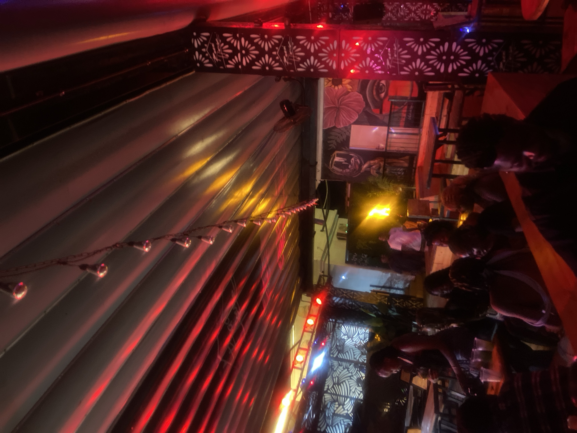 The image depicts a dimly lit outdoor gathering at night, likely a bar or restaurant, with patrons seated at tables. The environment features a covered ceiling with rows of lights casting red and yellow hues. Decorative metal pillars with floral cutouts are visible, along with murals on the walls. Several people are present, some engaged in conversation or enjoying drinks. The overall atmosphere appears social and relaxed. The location is identified as Howayi, Nigeria.