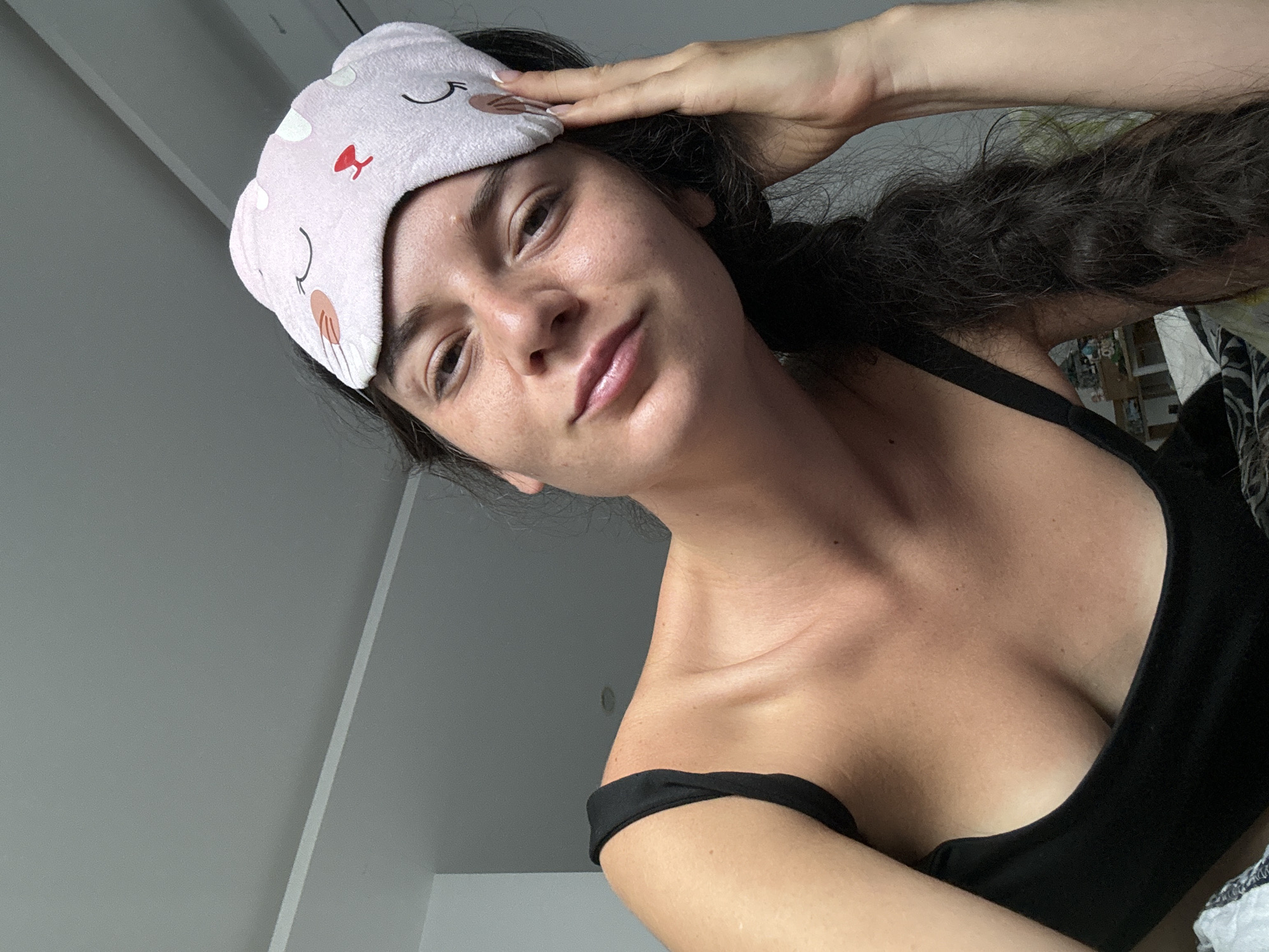 A young woman with dark hair braided and a pink sleep mask on her head, is lying in bed. She is wearing a black tank top. Her left hand is raised to her head, touching the sleep mask. The room appears to be a bedroom, with a ceiling and a wall visible in the background. The lighting is natural and appears to be coming from a window. There are no specific indicators of the location being in Mũi Né, Vietnam.