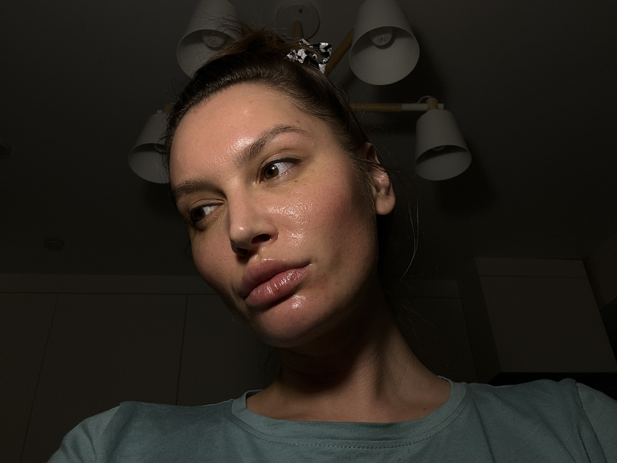 A low-angle close-up shows a woman with light skin, brown hair pulled back in a messy bun with a black and white patterned scrunchie, wearing a blue-green t-shirt. Her face appears shiny, possibly due to oil or moisturizer. She is looking off to the side. Above her is a modern ceiling light fixture with multiple white lamps. The background is dark and indistinct. There is nothing in the image to indicate the city of Moscow, Russia.