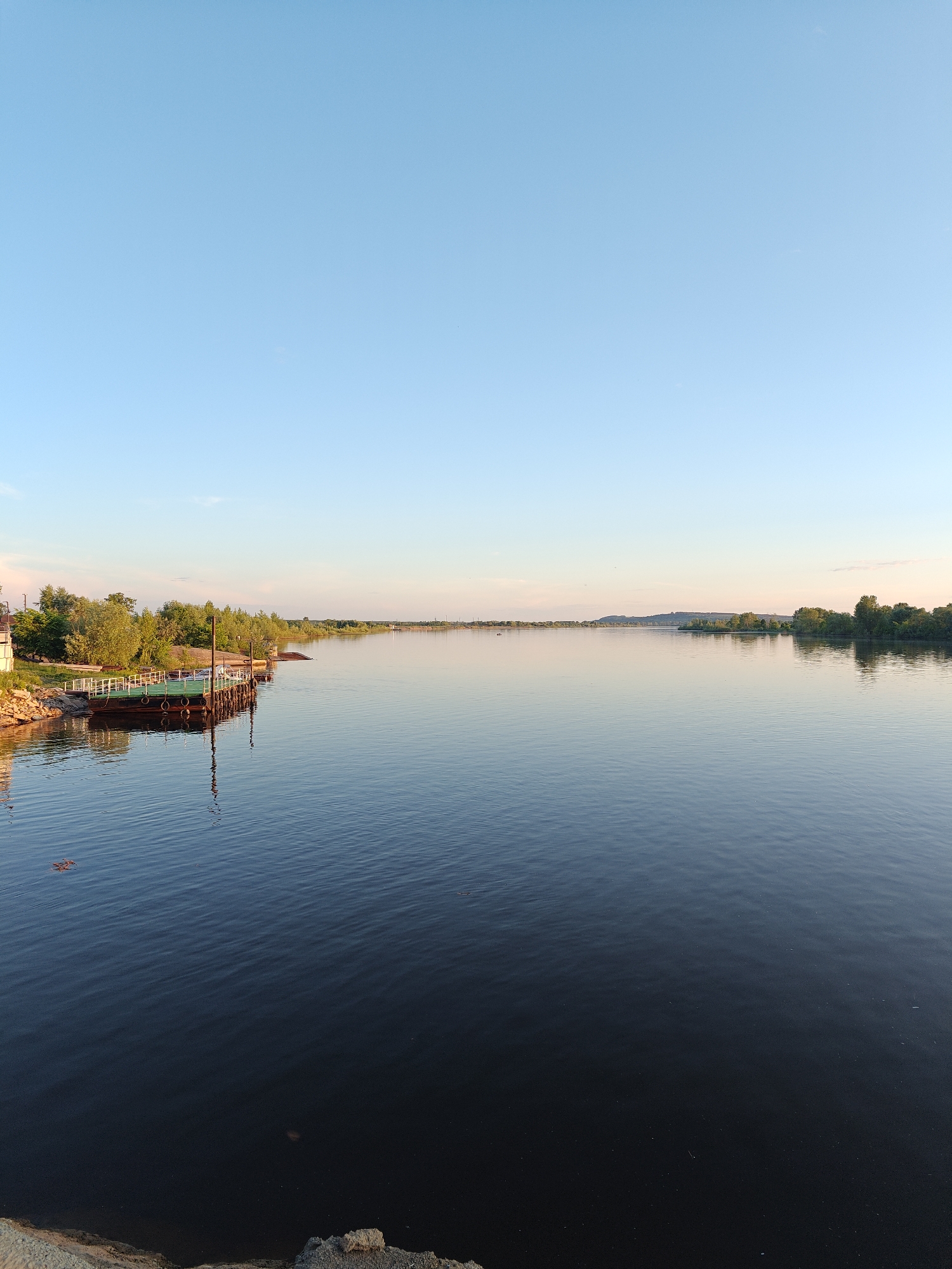 The image depicts a panoramic view of a wide, calm river at Nikolo-Beryozovka, Russia, under a clear, light blue sky during late afternoon or early evening. The water, a deep blue-green, occupies the majority of the lower frame, showing gentle ripples and reflections of the sky and distant banks.
On the left bank, a sturdy, flat-topped metal pier with a green deck is anchored, featuring multiple black rubber tires along its visible side, likely for cushioning. Behind the pier, dense green deciduous trees and foliage line the bank, illuminated by warm sunlight. Faint white structures, possibly railings or small buildings, are visible near the leftmost edge of the image on the bank. The immediate foreground on the left shows a rocky, earthen bank.
The river extends towards the horizon, where the distant right bank is lined with a continuous stretch of green trees and elevated terrain, possibly low hills or bluffs. The overall atmosphere is tranquil and devoid of human activity or explicit figures. The sky is predominantly clear, with minimal atmospheric haze near the distant horizon.