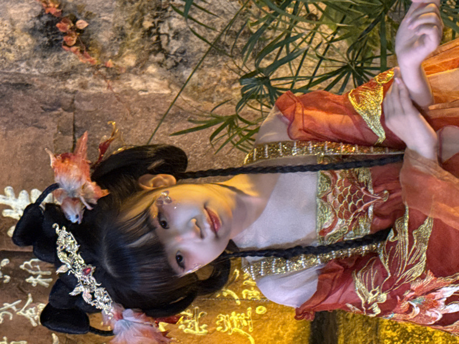 A young East Asian female, estimated to be a child, is depicted in a posed, illuminated shot. She wears elaborate traditional Chinese attire, featuring a multi-layered red and orange garment with extensive gold embroidery, including a prominent bird or phoenix motif on the chest, and gold beaded trim. Her hair is styled in two large black buns with long braids extending over her shoulders, adorned with a golden filigree headpiece containing a red jewel and decorative pink and orange fabric elements. Her face is made up with a light base, pink tones around the eyes, a soft red lip, and glitter or decorative elements applied below the left eye. Her head is slightly tilted, and she gazes directly at the viewer with a subtle smile. Her hands are gently crossed in the lower right of the frame. The background to the left features a stone wall with partially visible golden Chinese characters, including "金" (jīn) and "柳" (liǔ) or "柿" (shì), suggesting a cultural or historical setting within the city of Yuzhong District, China. To the right, green leafy plants and darker rock textures are visible. The scene is illuminated by warm, artificial light, creating a golden glow.