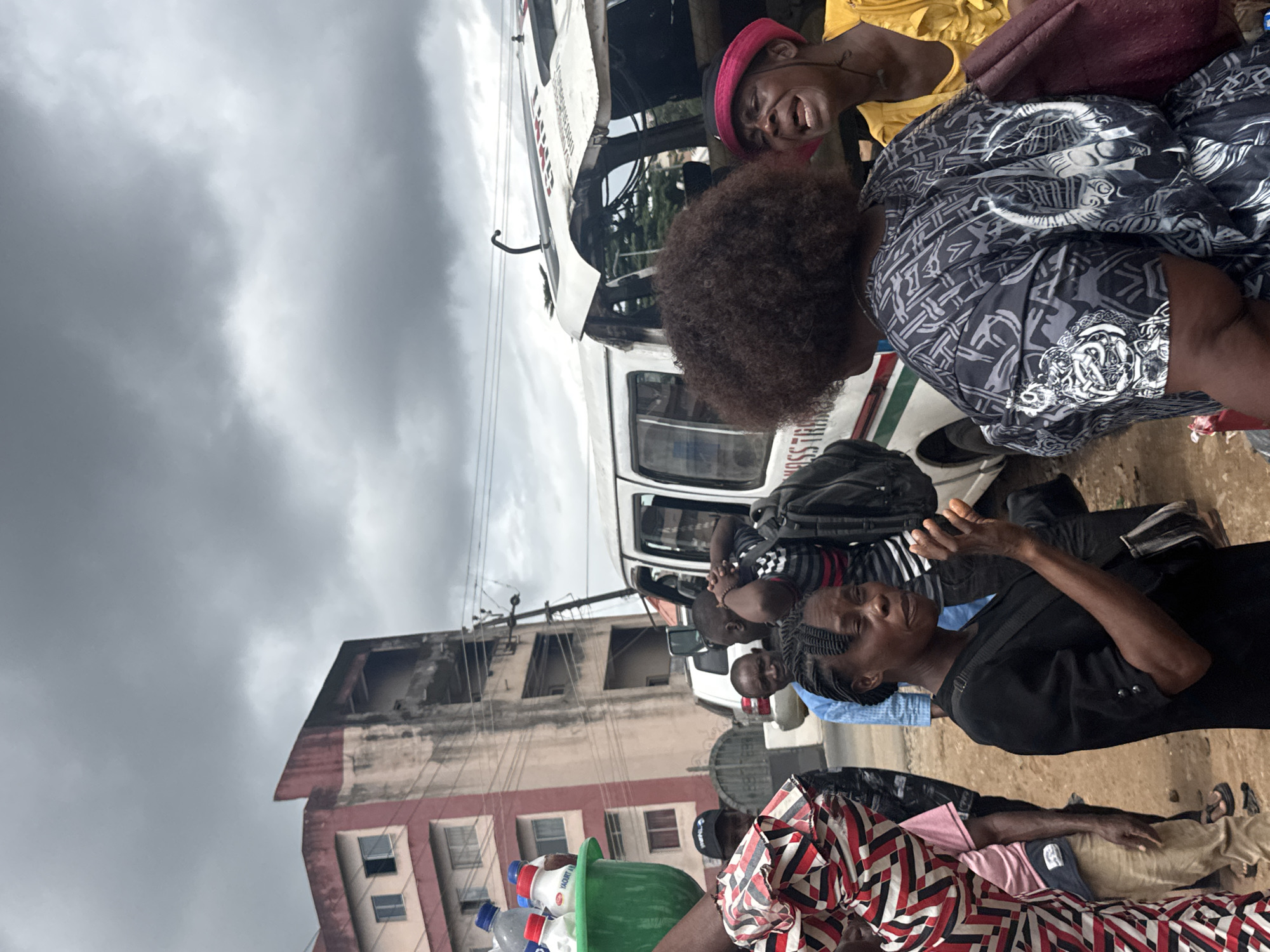 In Owerri, Nigeria, on a cloudy day, a scene unfolds with an overturned white passenger bus displaying "PASS TRANS" on its side, suggesting a transportation company. Several people are gathered around the bus, some appearing distressed. A woman in the foreground, wearing a black dress and a black and white patterned bag, gestures with her right hand, her mouth open in what seems to be an expression of concern or explanation. To her right, another woman with a bright pink headscarf and a yellow top is visibly upset, her mouth agape and eyes closed, as if crying out. Behind them, more individuals are present, including a man in a blue shirt and others near the damaged bus. In the background, a multi-story building with a faded pink and concrete facade stands, its upper floors appearing aged and possibly abandoned. Bottles of water and what appears to be yogurt are visible in a green container in the left foreground, hinting at goods being transported or sold. The ground is dirt and gravel, indicating an outdoor, possibly roadside location. The overall atmosphere suggests a recent incident, possibly an accident, and the emotional responses of the individuals point to a significant event.
