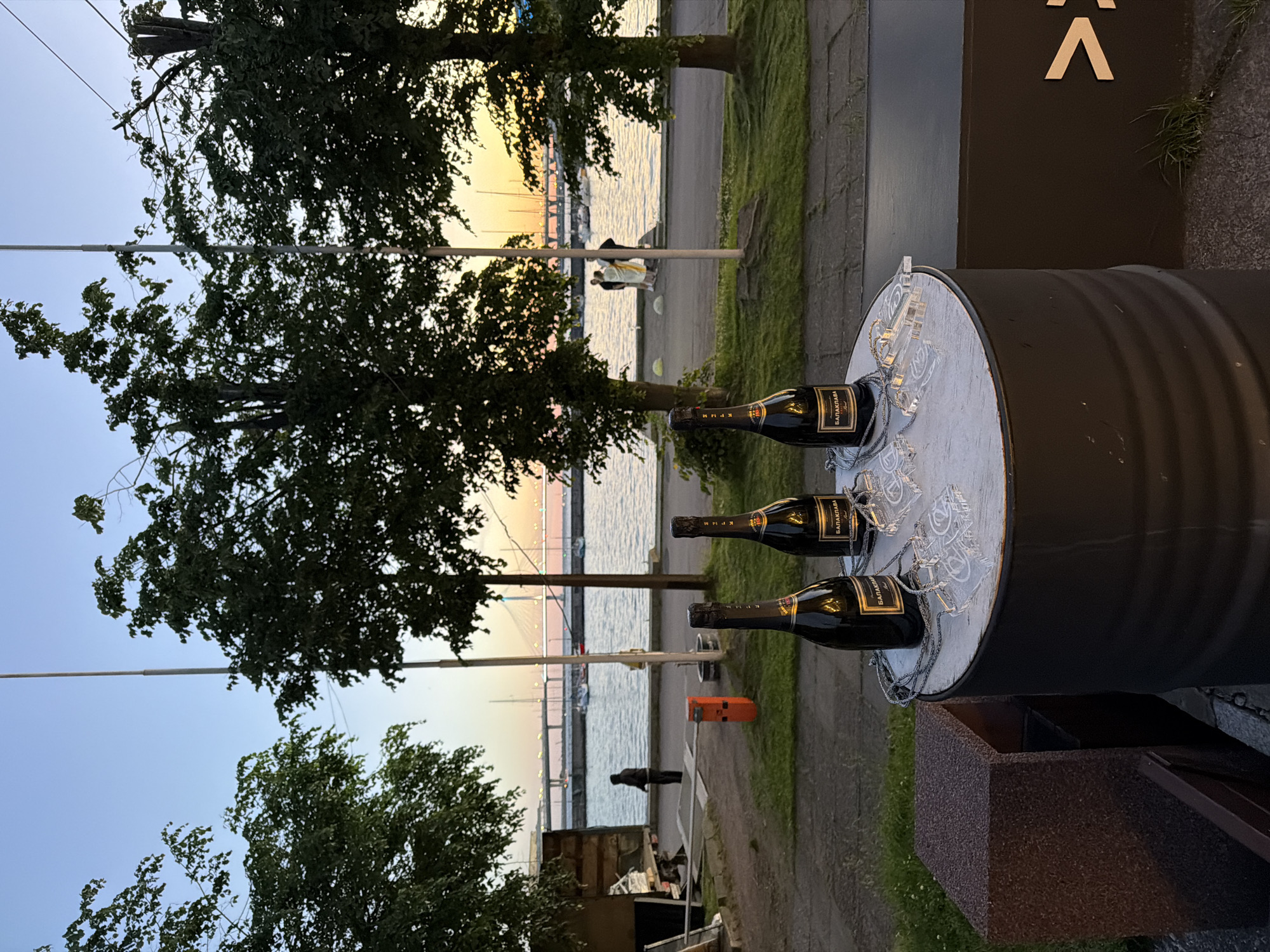 At sunset, three bottles of sparkling wine, identified as "BALAKLAVA," are arranged on a round table made from a repurposed industrial barrel. Several clear, flat objects, possibly coasters or decorative elements, are scattered on the table, some appearing to have numbers or symbols etched onto them. In the background, a body of water, likely a river or bay, is visible, with sailboats and a bridge in the distance. The scene is framed by lush green trees. A person is standing by a metal structure on the left, and a few individuals are gathered near the water's edge in the middle ground. The lighting suggests it is late evening, with the sky displaying hues of orange and pink from the setting sun. The location is St. Petersburg, Russia, with the architecture of the bridge and the presence of sailboats being characteristic of the city's waterfront areas.