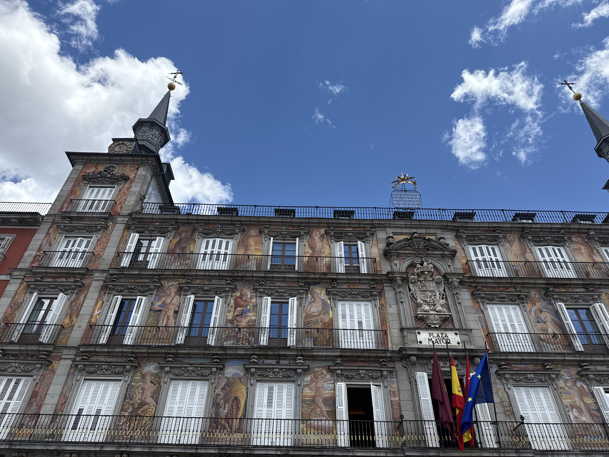 The image shows a building in Plaza Mayor, Madrid, Spain. The building has multiple floors with windows and balconies. The facade is decorated with paintings and sculptures. Several flags, including the Spanish flag and the European Union flag, hang from a balcony in the center of the building. The sky above is blue with scattered clouds. There are two towers with spires visible on the top corners of the building.