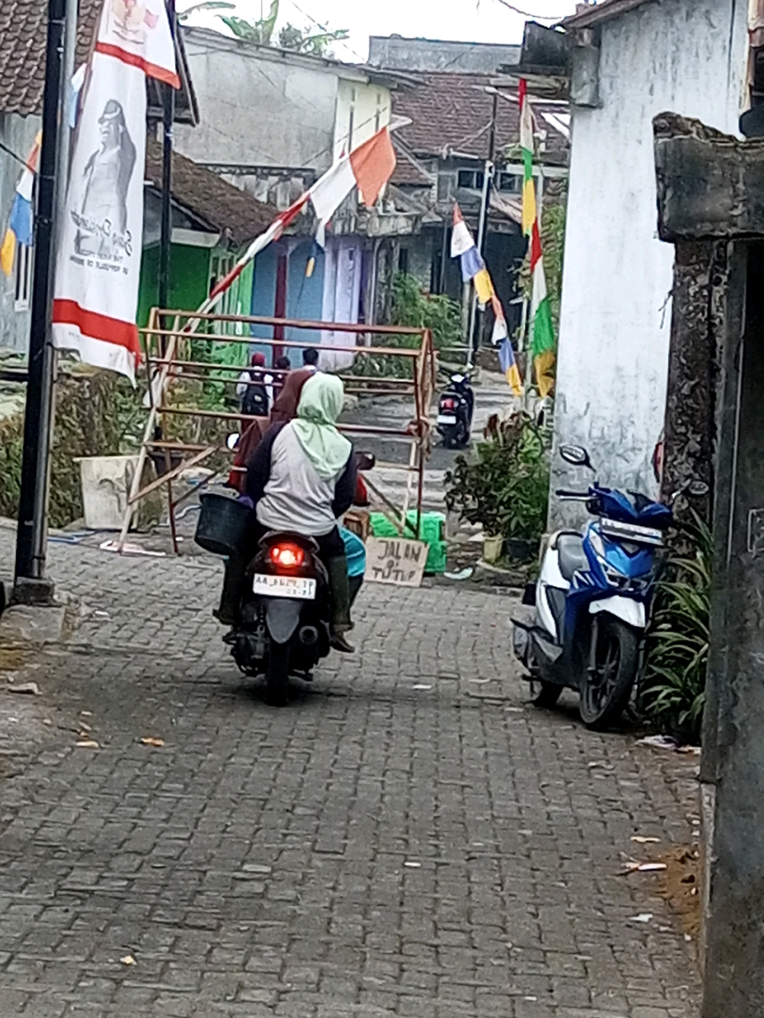In Andongsili, Indonesia, a person wearing a light green headscarf and a dark long-sleeved shirt rides on the back of a scooter. They are seated behind the driver and appear to be carrying a black bucket. The scooter has a license plate with the characters "AA 3629 ZP". The scooter is on a cobblestone street. In the background, there are several houses with tiled roofs, some of which are brightly colored. Flags are visible, including a large banner with a black and white image of a person. Another scooter is parked on the side of the street. A sign on the left reads "JALAN TUTUP", which translates to "Street Closed". The scene depicts a typical street view in a residential area, possibly during an event or a day where certain roads are temporarily closed.