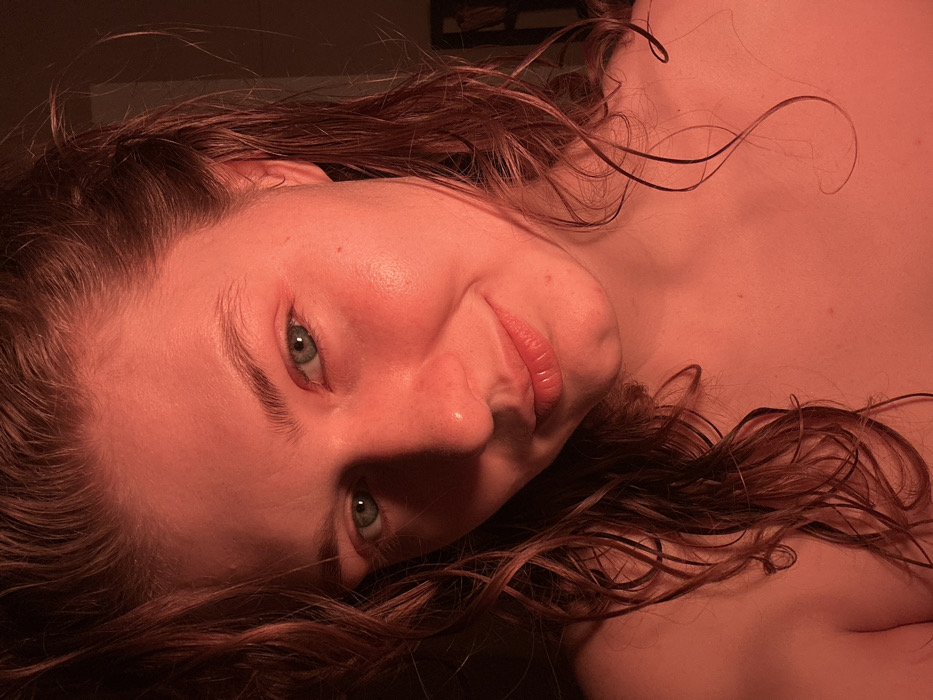 A close-up, chest-up portrait depicts a female subject with light skin, wet, wavy reddish-brown hair, and green eyes, illuminated by strong red light. Her face is turned slightly to the right, and she looks directly at the camera with a subtle smile. Her shoulders are bare. The background is dark and out of focus, suggesting an indoor setting. The scene is located in the city of Kazan, Russia.