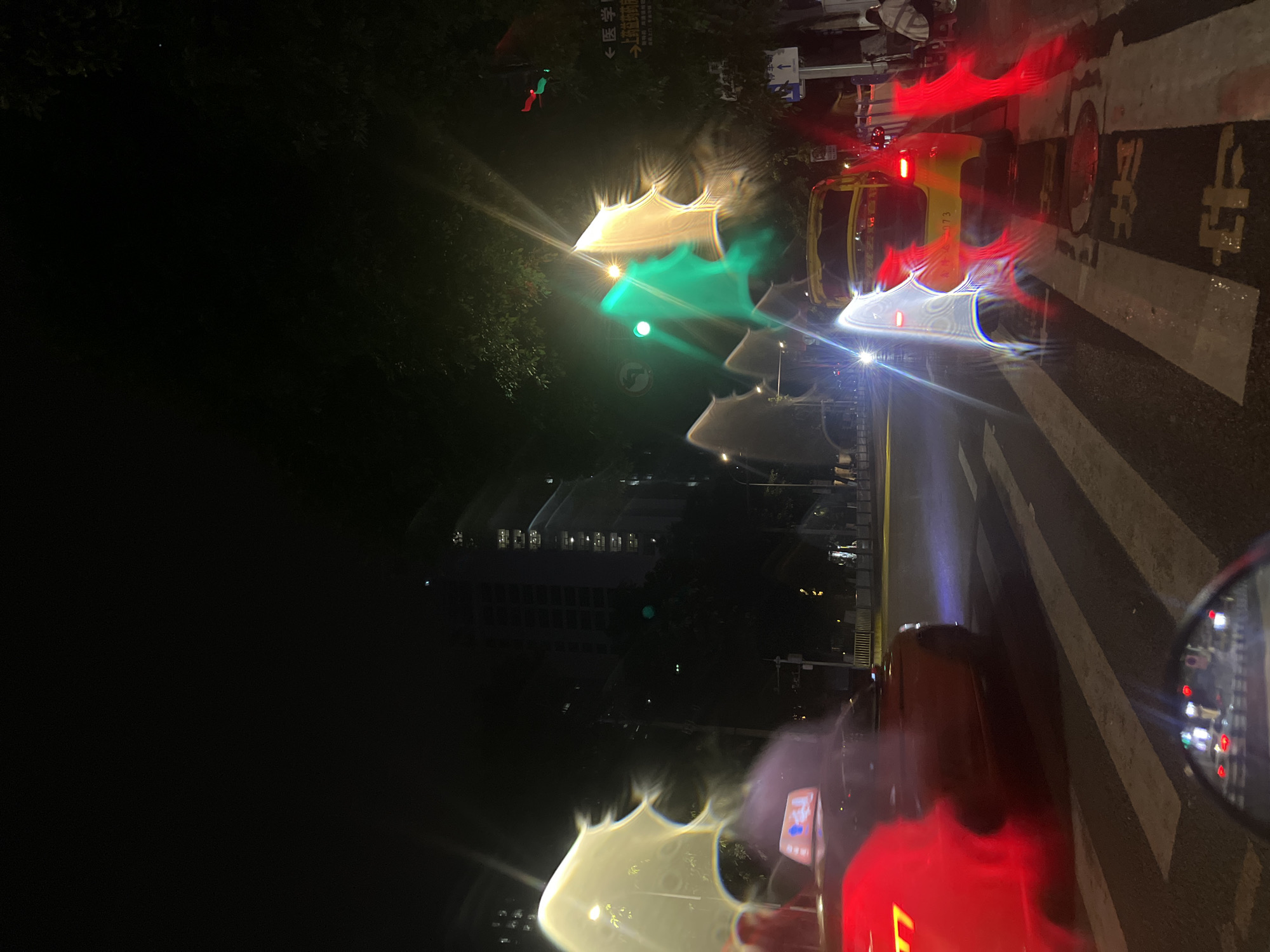 This is a night time photograph taken from a moving vehicle, likely a motorcycle, in the Jiulongpo District of China. The image captures a street scene with several taxis. In the foreground, a red taxi is visible on the left, its taillights illuminated. To the right, a yellow taxi is parked on a crosswalk, its rear visible. The street is wet, reflecting the lights of the vehicles and traffic signals. Traffic lights are visible, with a green light illuminated. Buildings can be seen in the background, suggesting an urban environment. The lighting conditions are low, and there are significant light flares and reflections, likely due to water on the camera lens. The overall impression is that of a busy street at night, with the motion blur indicating the photographer is also in transit.